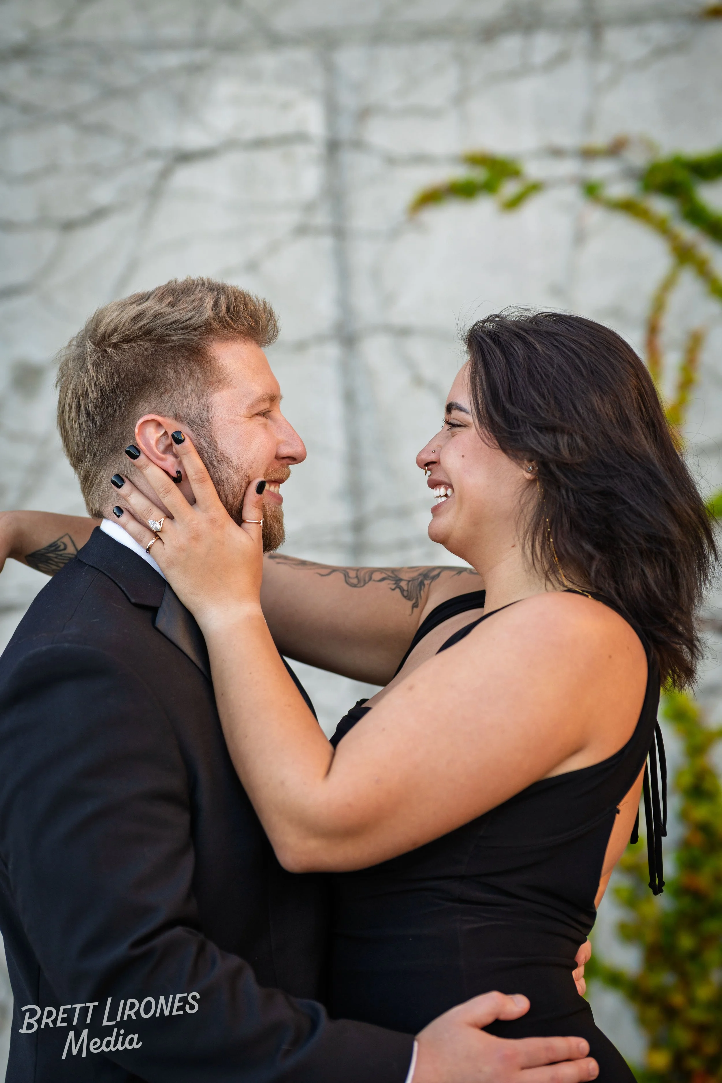 A man and woman share a joyful moment, facing each other closely and smiling. The woman holds the man's face with both hands. They appear to be happy and in love, with a plain, light-colored background and some greenery in the scene.