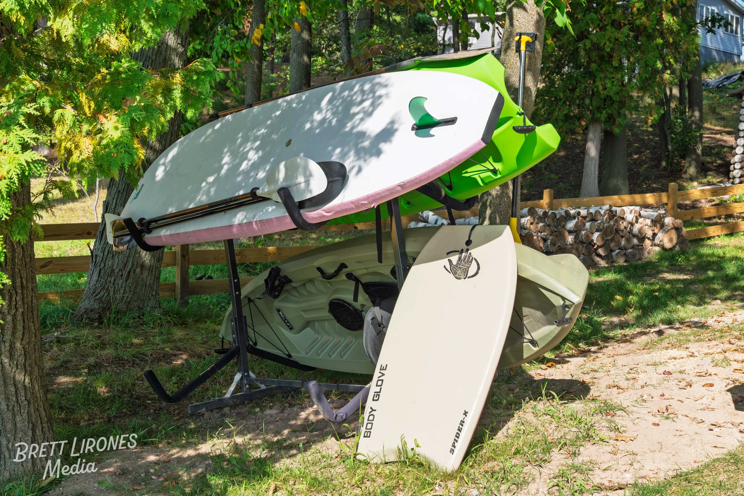 A kayak and paddleboard are stacked upside down beneath a tree outdoors, with a wooden fence and firewood in the background.