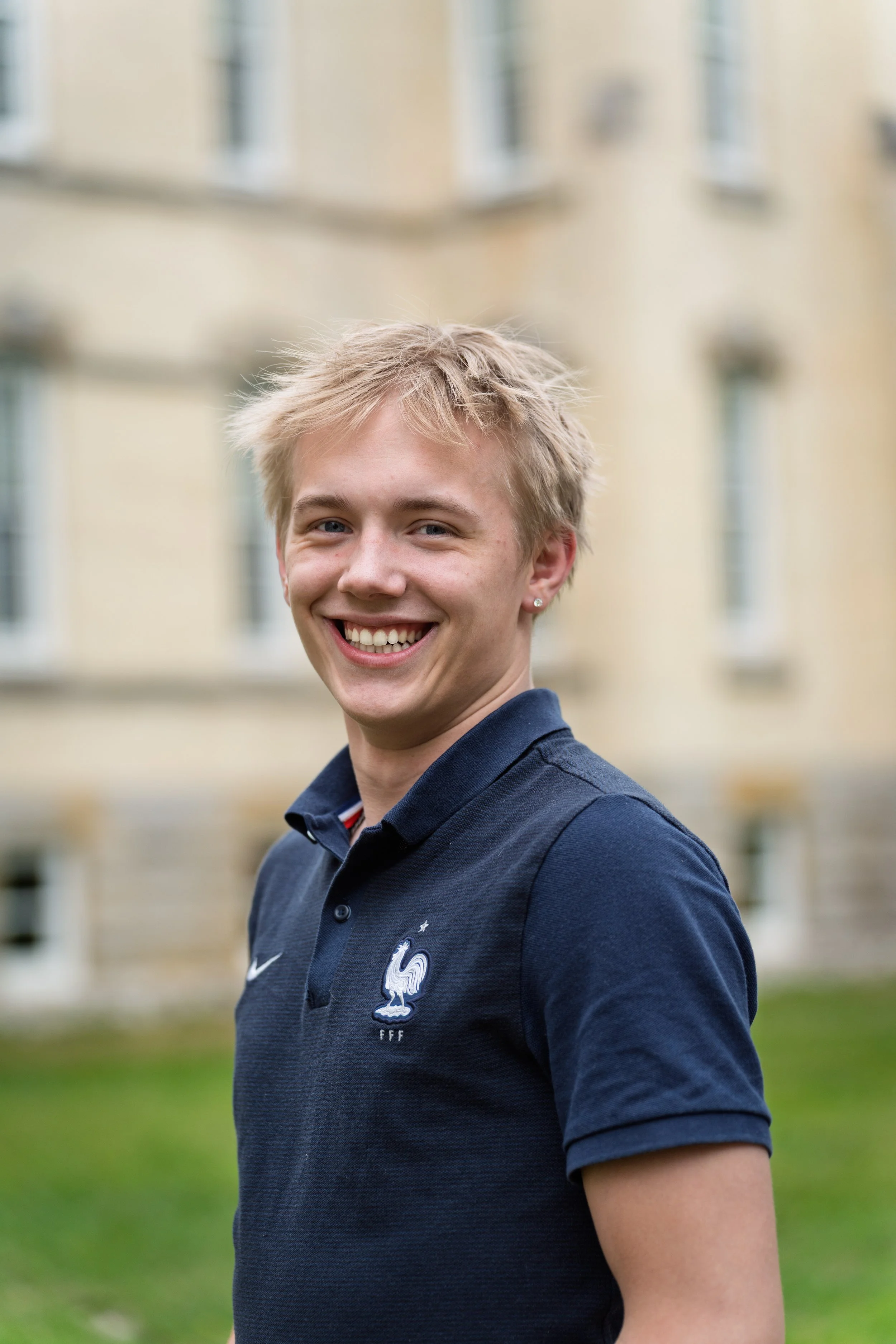 A young man with short blonde hair, smiling, outside in front of a blurred building, wearing a navy blue polo shirt with a French football team logo.