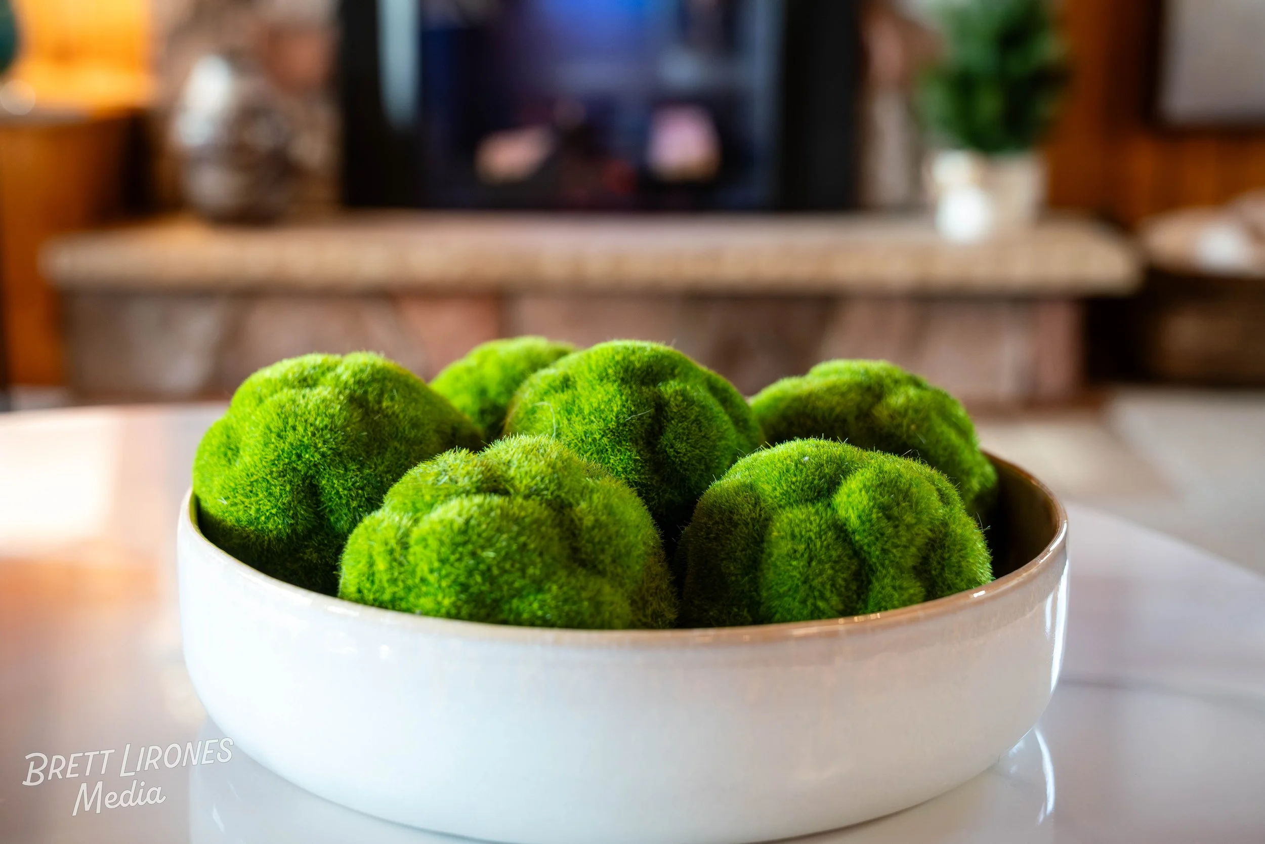 A white bowl filled with green moss balls placed on a white surface in a cozy living room.