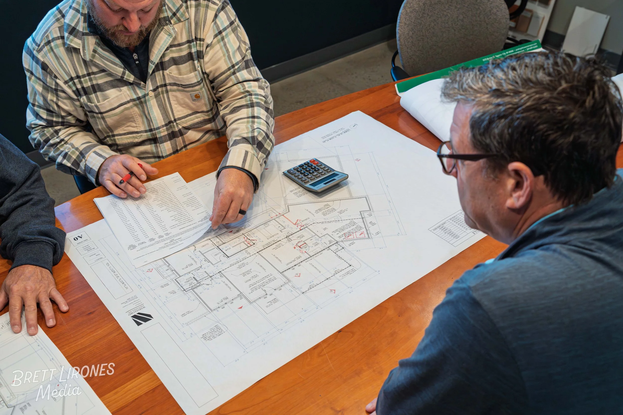 Two men discussing architectural blueprints and plans on a wooden table, with one holding a sheet of paper, a calculator, and other paperwork on the table.