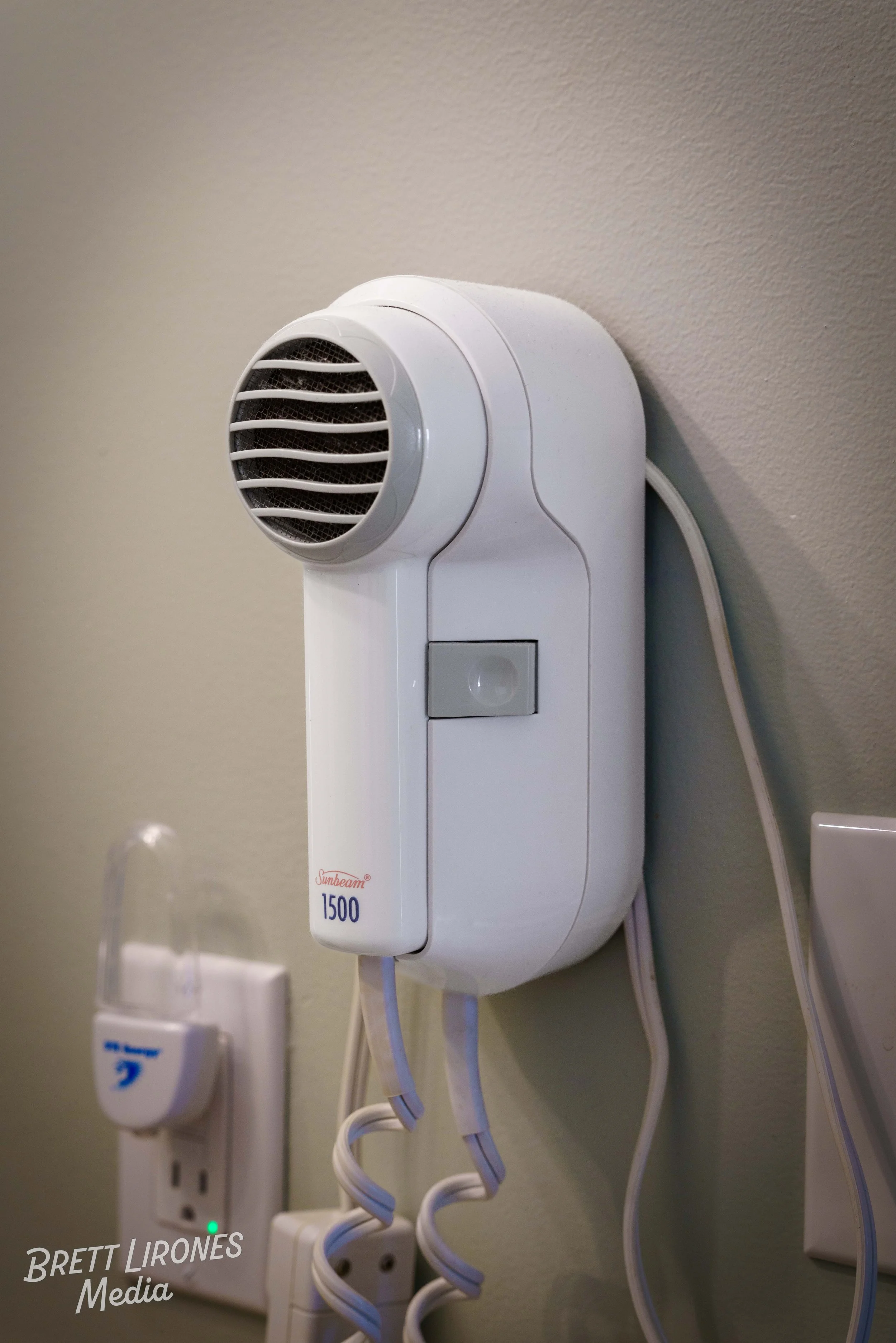 A white Sunbeam 1500 halo therapy device with a circular vent on top, plugged into an electrical outlet on a beige wall, with cords hanging down.