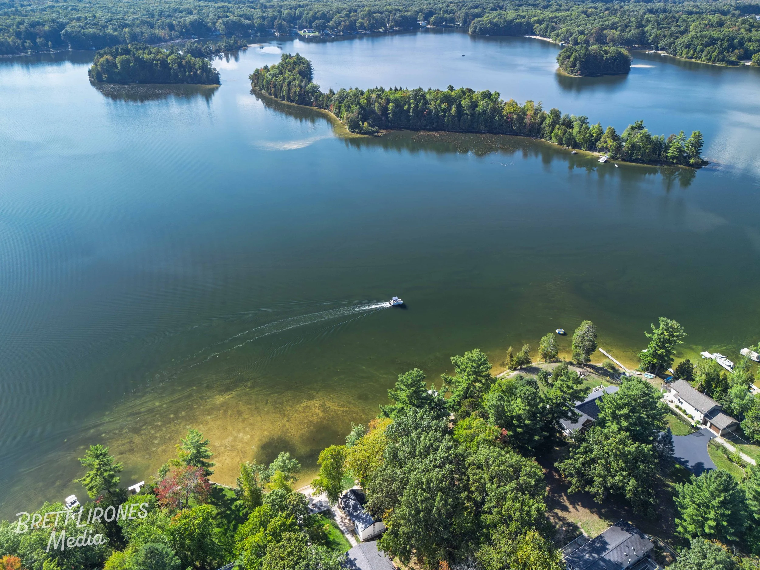 Aerial view of a lake with several small islands and surrounding forests, with a boat creating ripples on the water, and a shoreline with houses and trees.