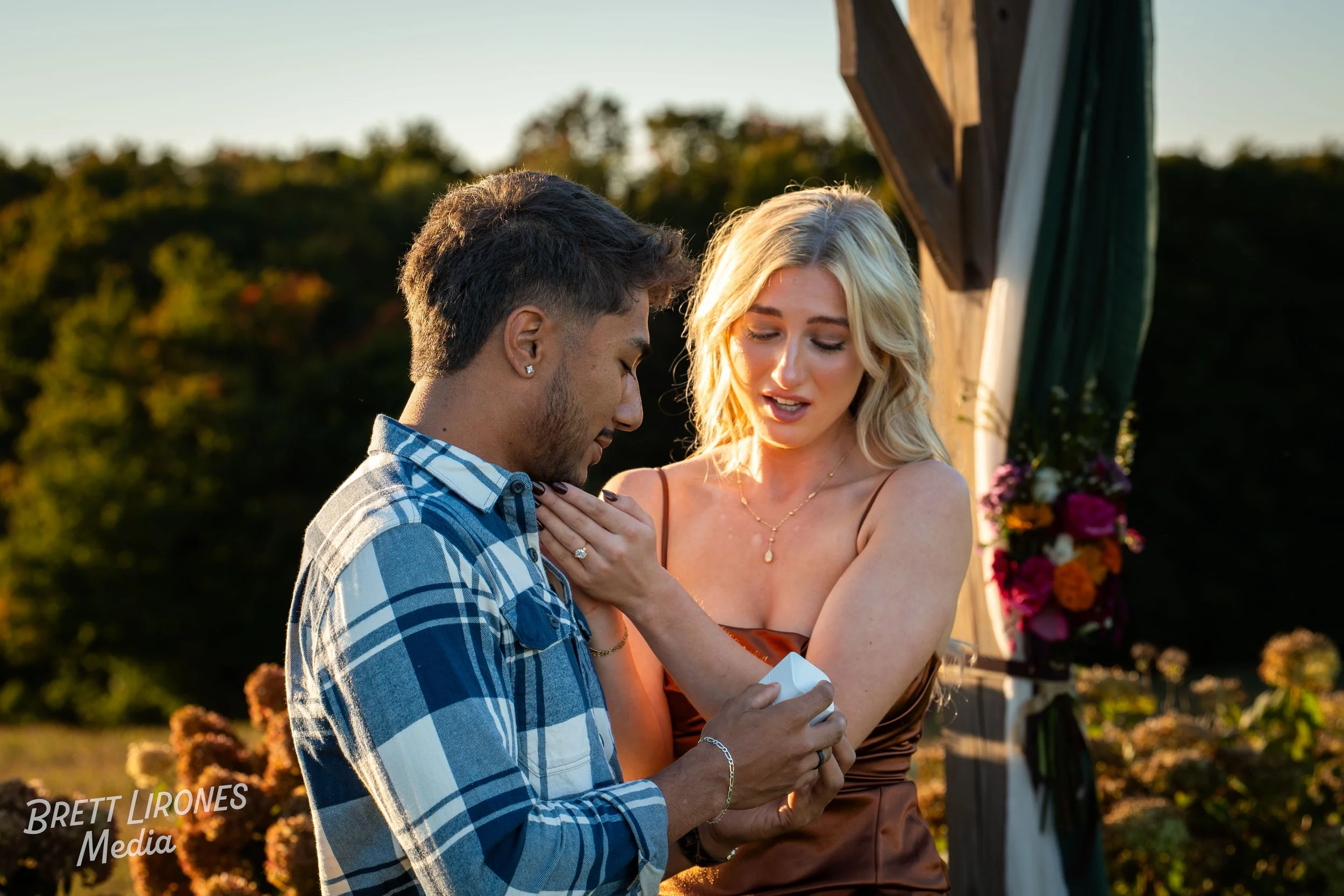 A couple during a wedding ceremony outdoors at sunset, with the woman placing a ring on the man's finger, in front of a wedding arch decorated with flowers.