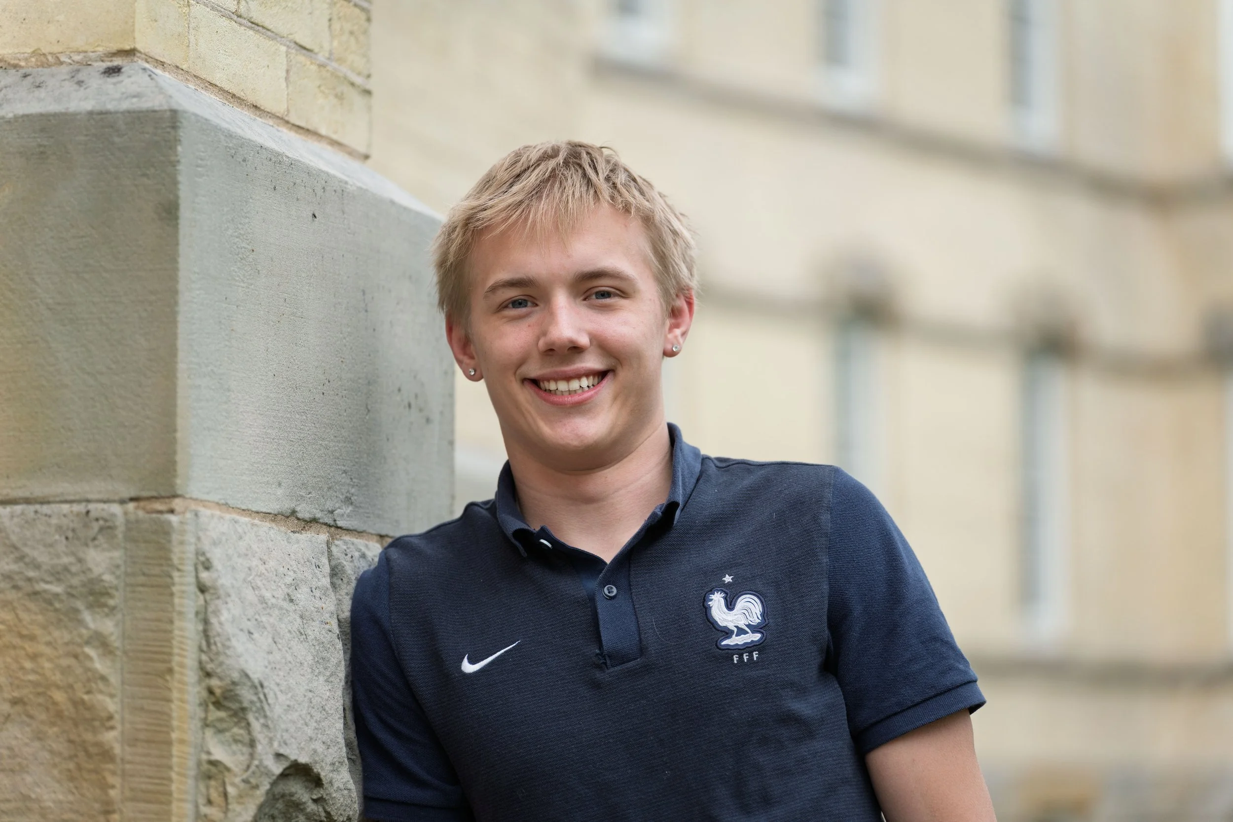 A young man with short blond hair, wearing a navy blue polo shirt with a French football team emblem, stands outside near a brick building and smiles at the camera.