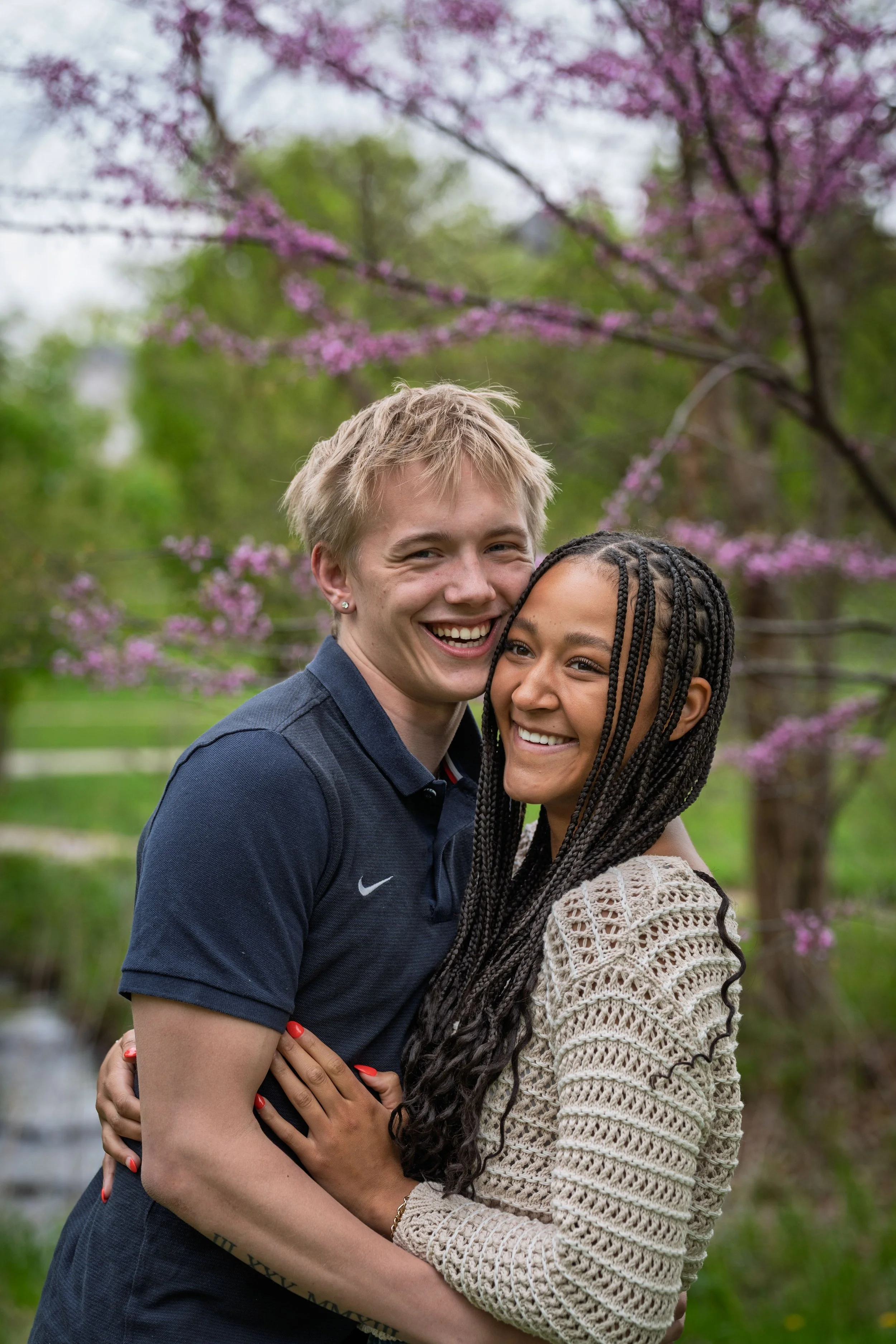 A smiling young couple hugging outdoors, with pink blossoms and green trees in the background.