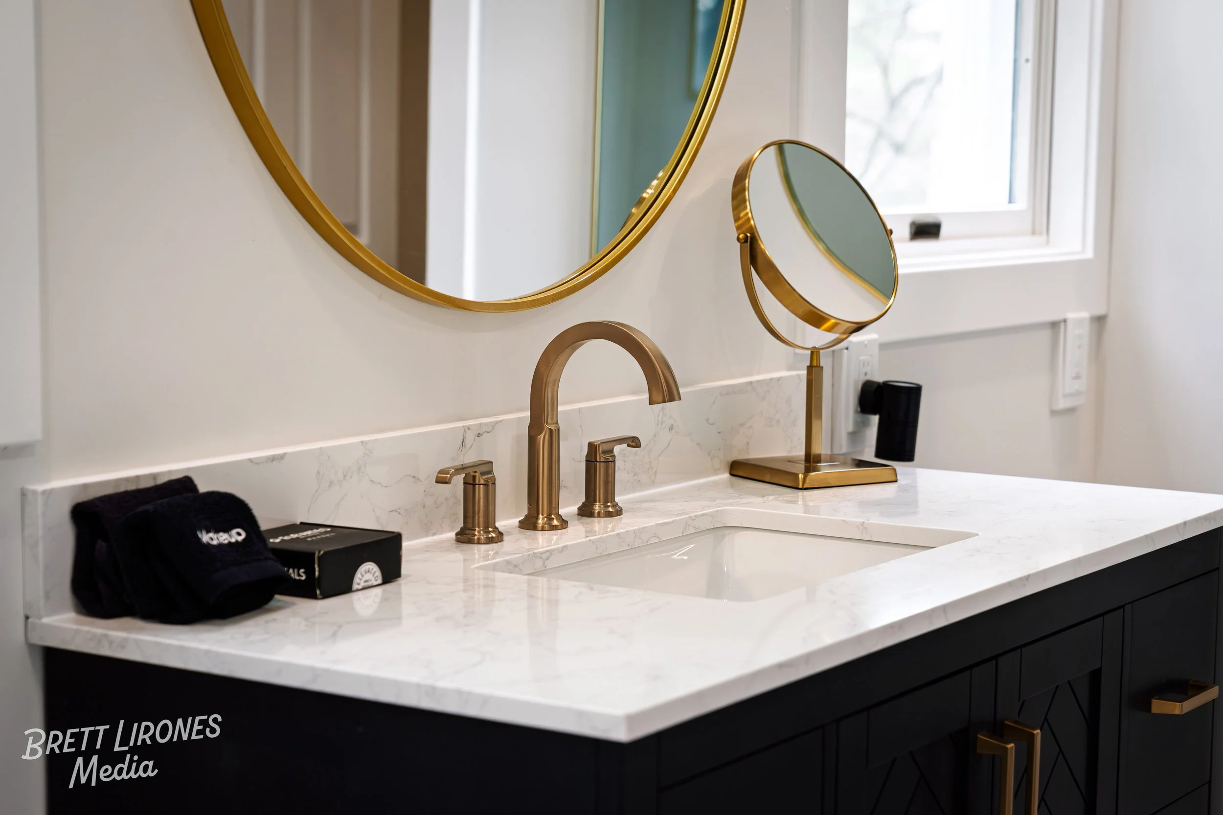 A bathroom sink with gold fixtures, a large round mirror with a gold frame, a smaller magnifying mirror, a plant in the background, and a window letting in natural light.