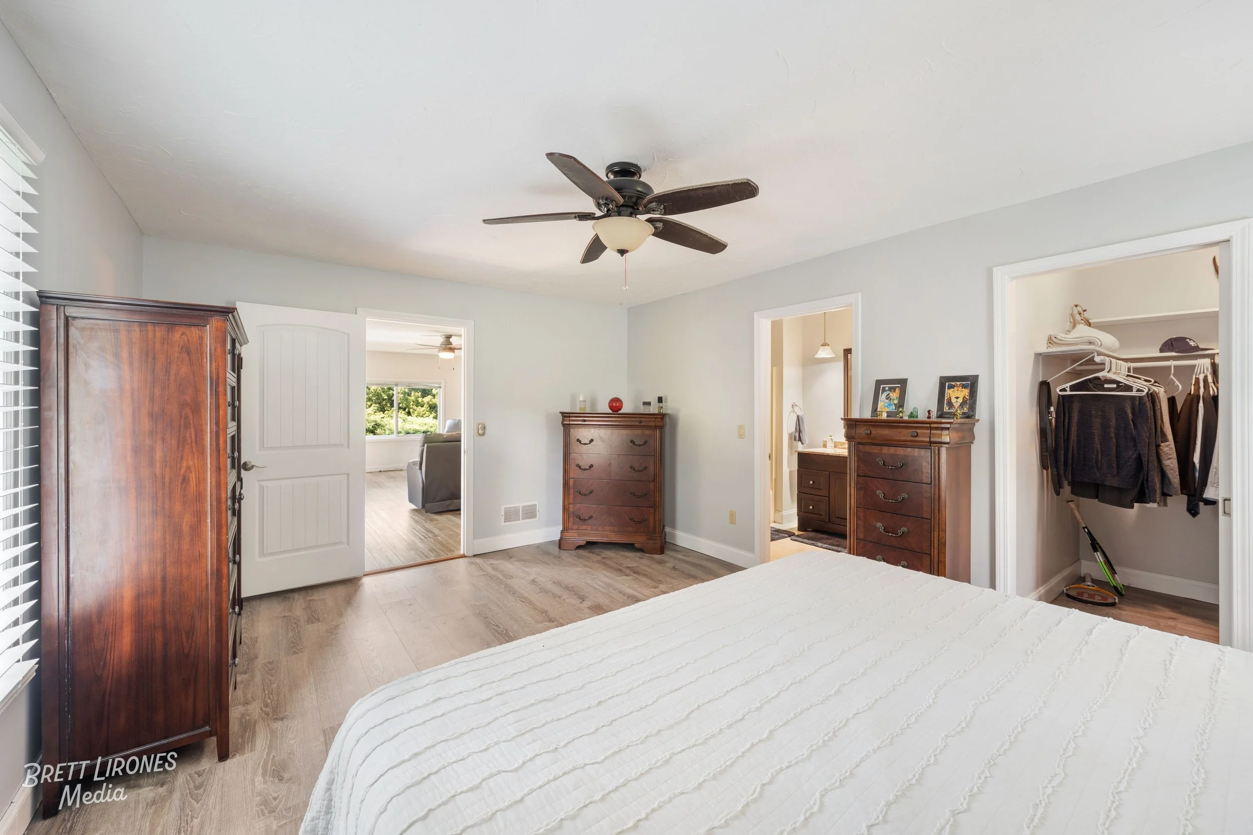 A bedroom with a ceiling fan, wooden dressers, and an open closet with clothes hanging inside. The bed has a white textured comforter, and there is a view into an adjoining room with a window and sofa.