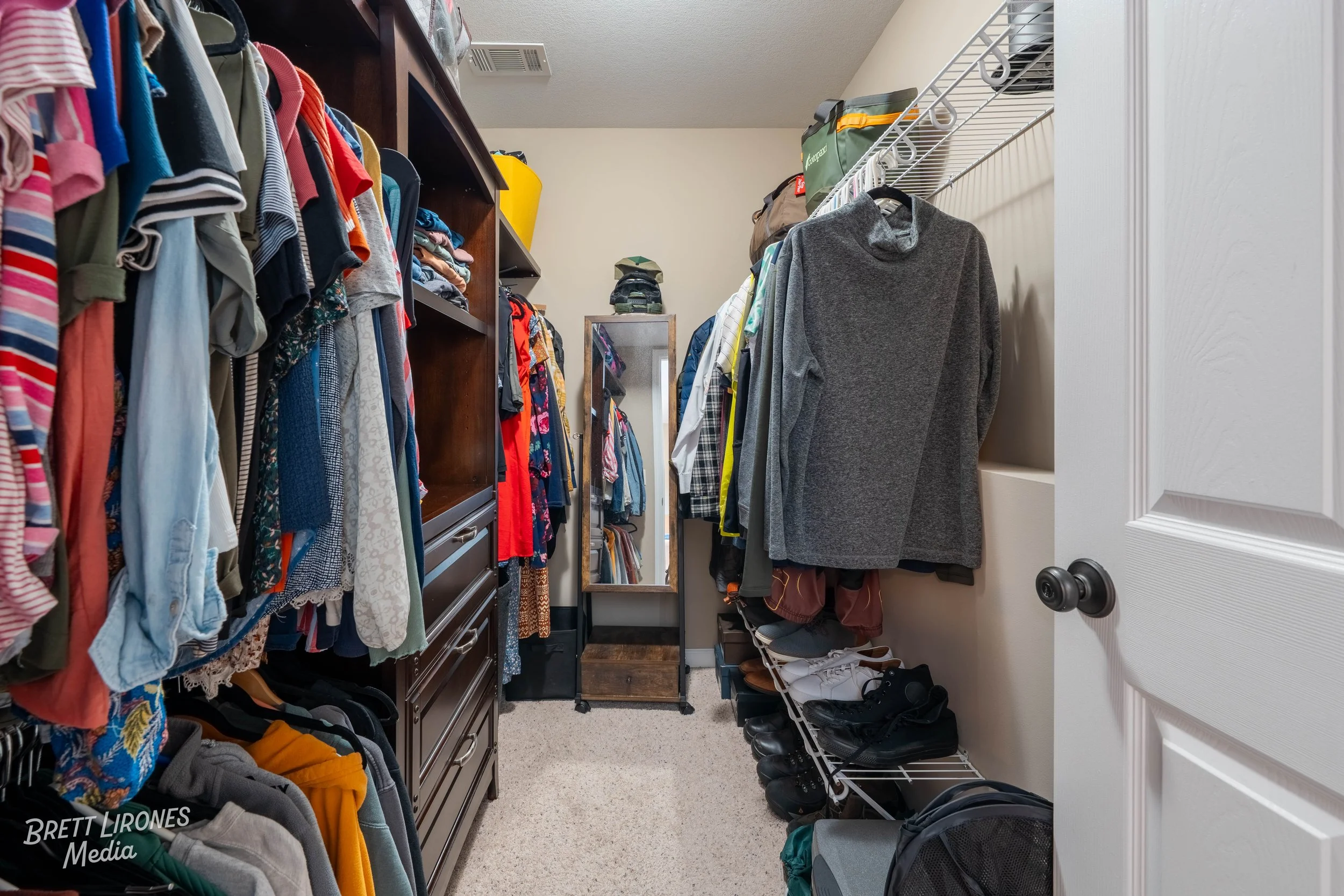 A cluttered walk-in closet filled with hanging shirts, folded clothes on shelves, shoes on a rack, and a full-length mirror at the back.