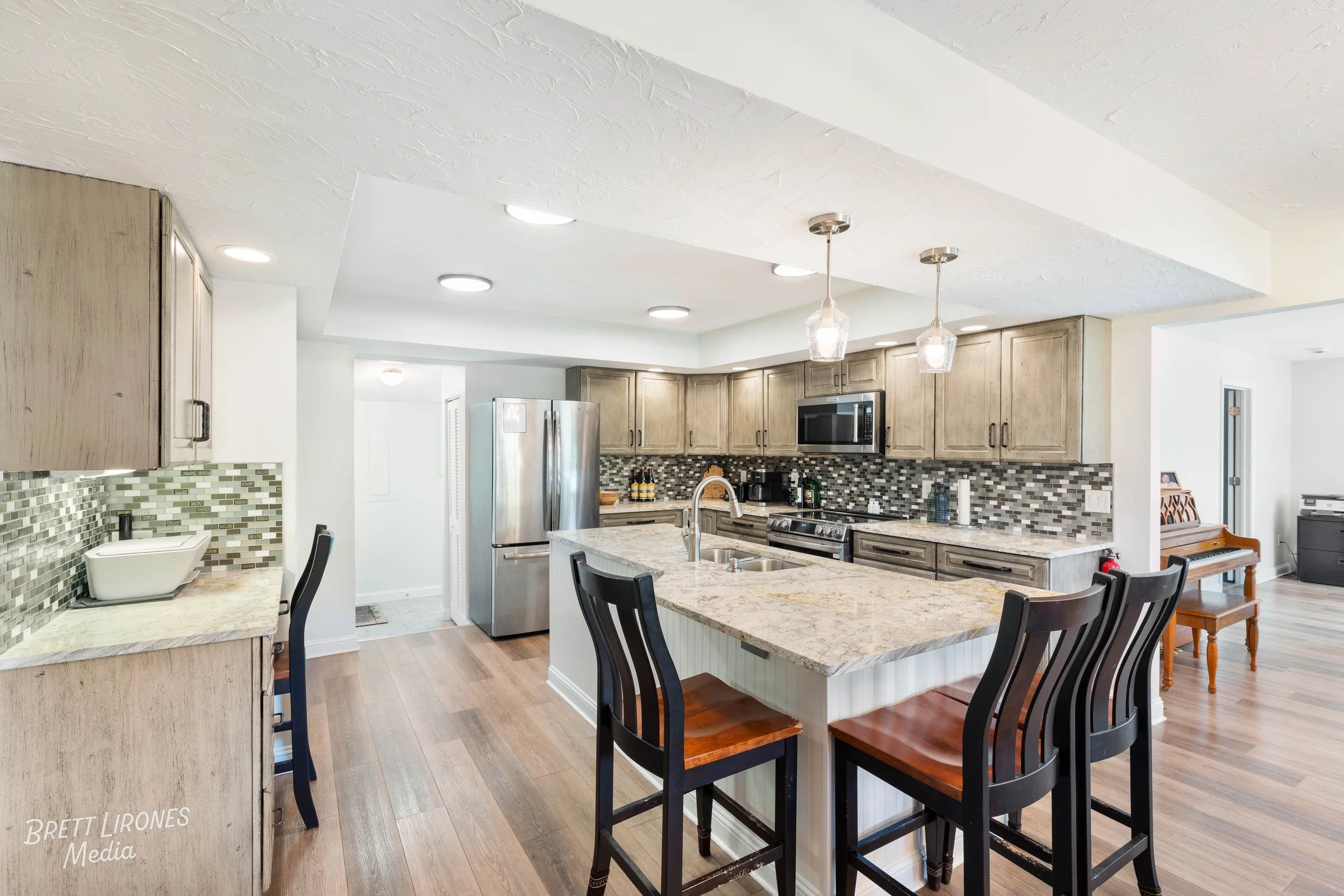 Modern kitchen with wood cabinetry, granite countertops, mosaic tile backsplash, stainless steel appliances, and a central island with seating. There are three black chairs with wooden seats at the island, pendant lights hanging above, and a dining a
