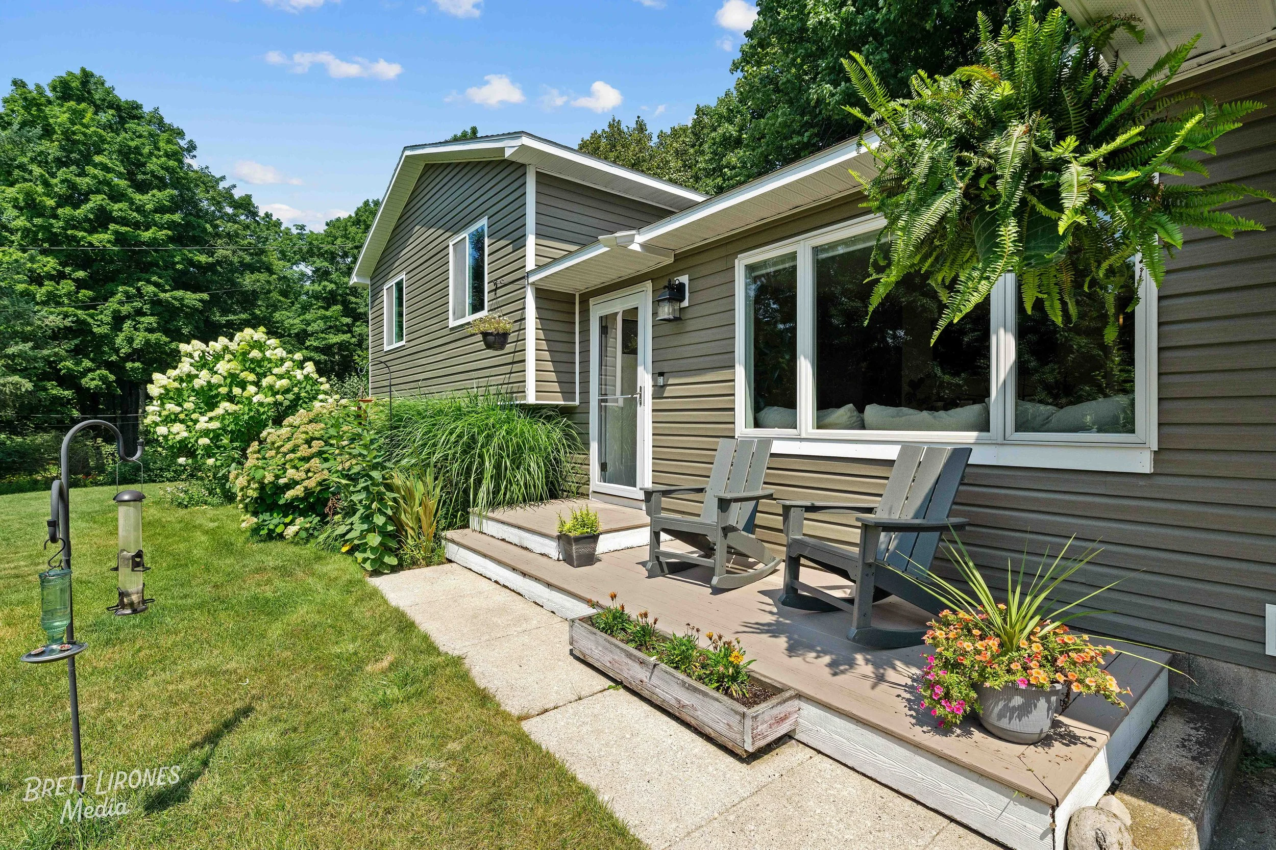 Backyard patio with wooden deck, two Adirondack chairs, potted plants, garden shrubs, and a screened door leading into a house, with a lush green lawn and trees in the background.