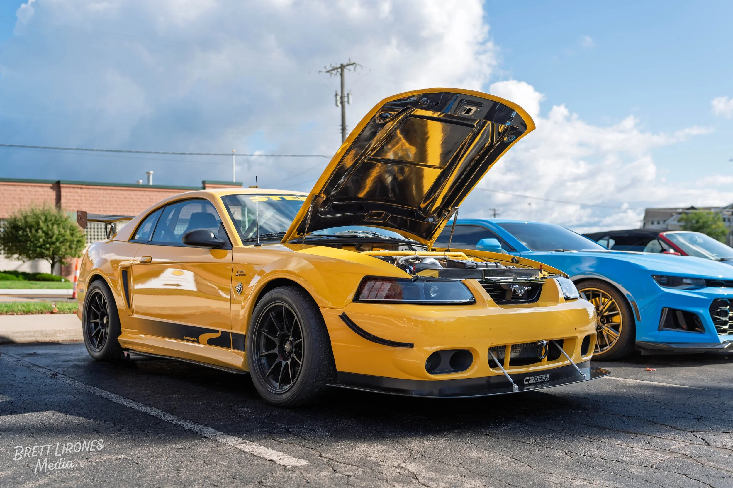 Yellow Ford Mustang with hood open, showing engine, parked beside a blue car, in a parking lot outdoors.