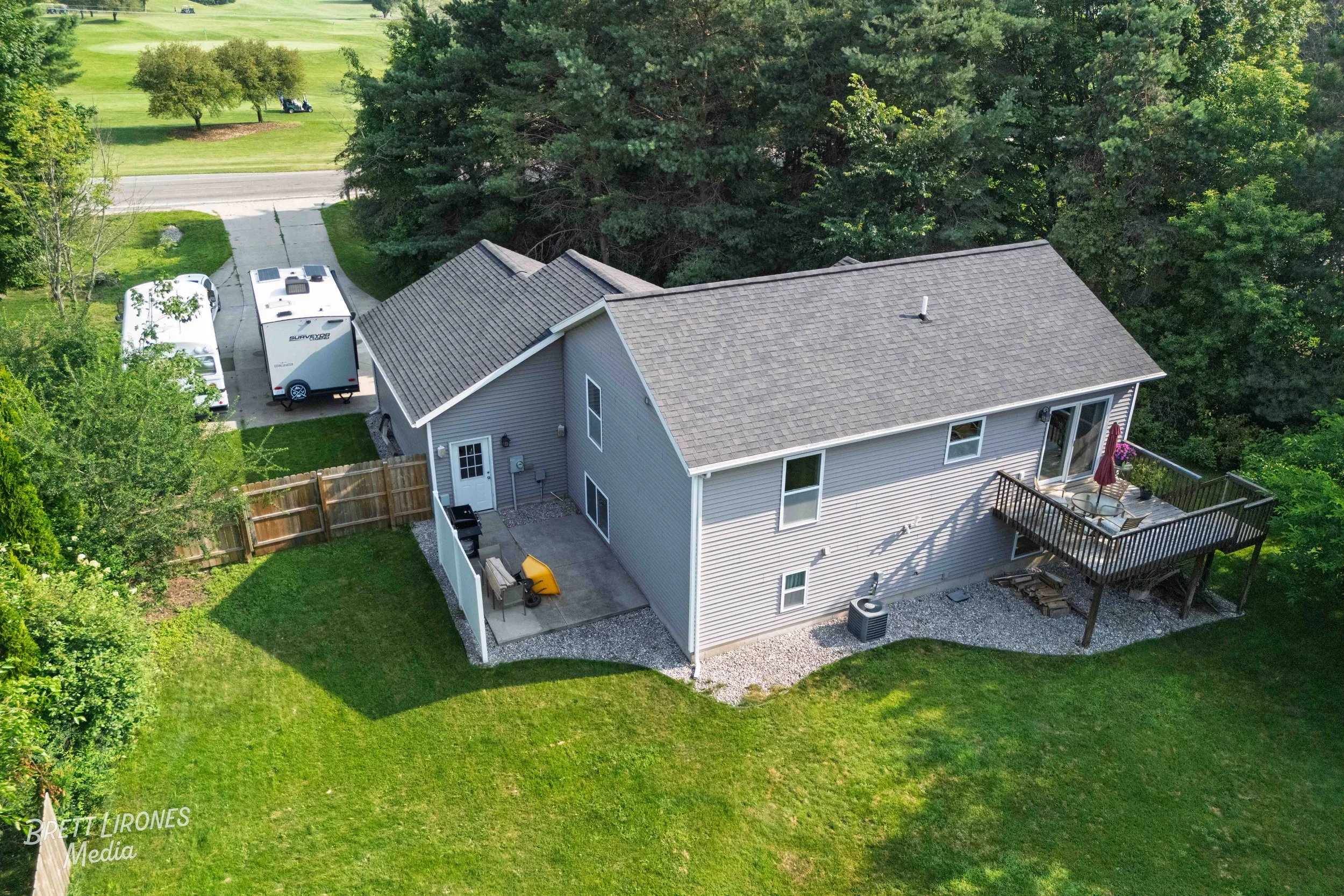 A two-story house with a large balcony overlooking a backyard with green lawn and trees. There are two recreational vehicles parked on the driveway, a small patio with outdoor furniture, and a fenced area near the house. The backyard is bordered by a