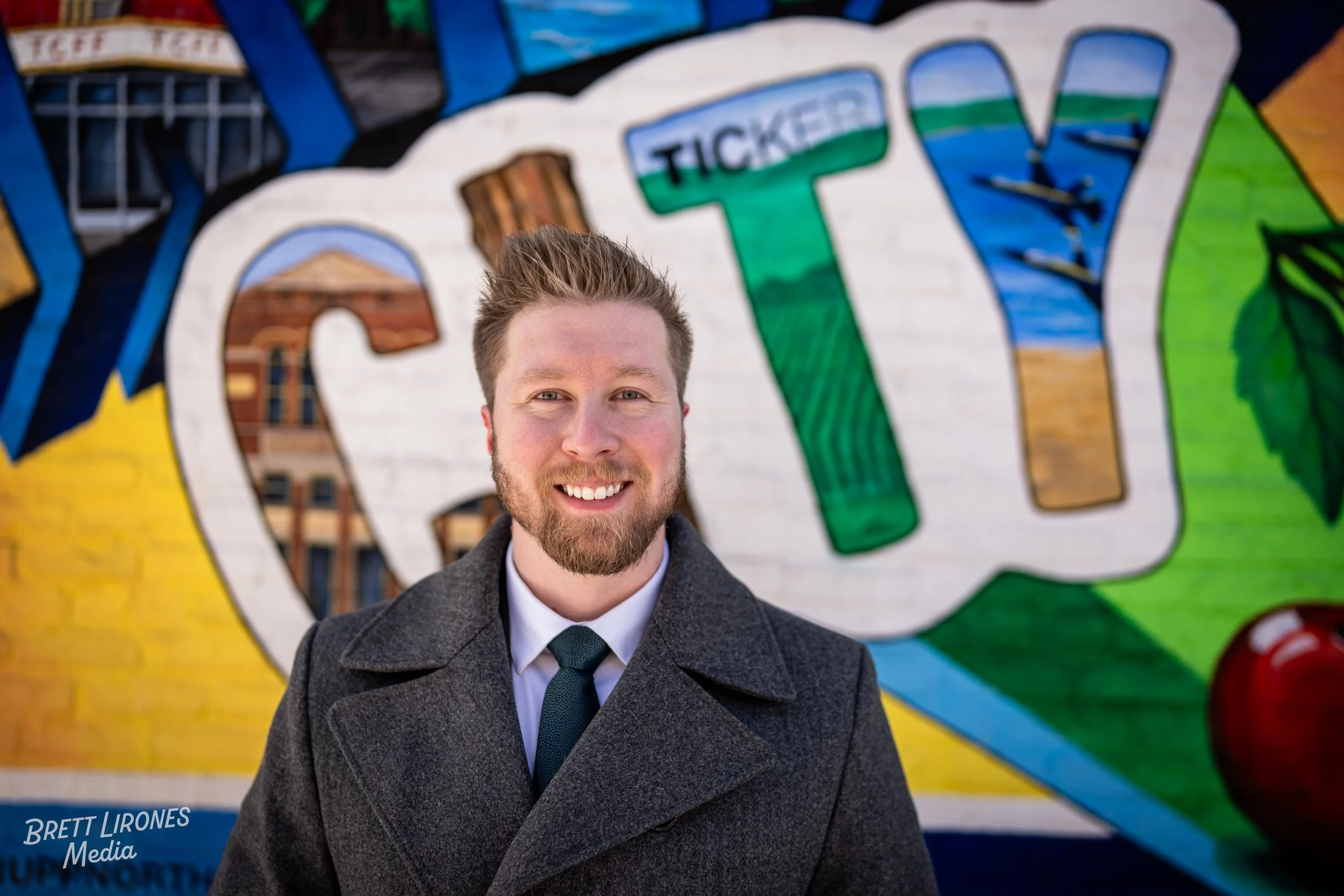A smiling man in a gray coat and tie stands in front of a colorful mural with the word 'City' painted on it, including the part 'TICKER' in the background.