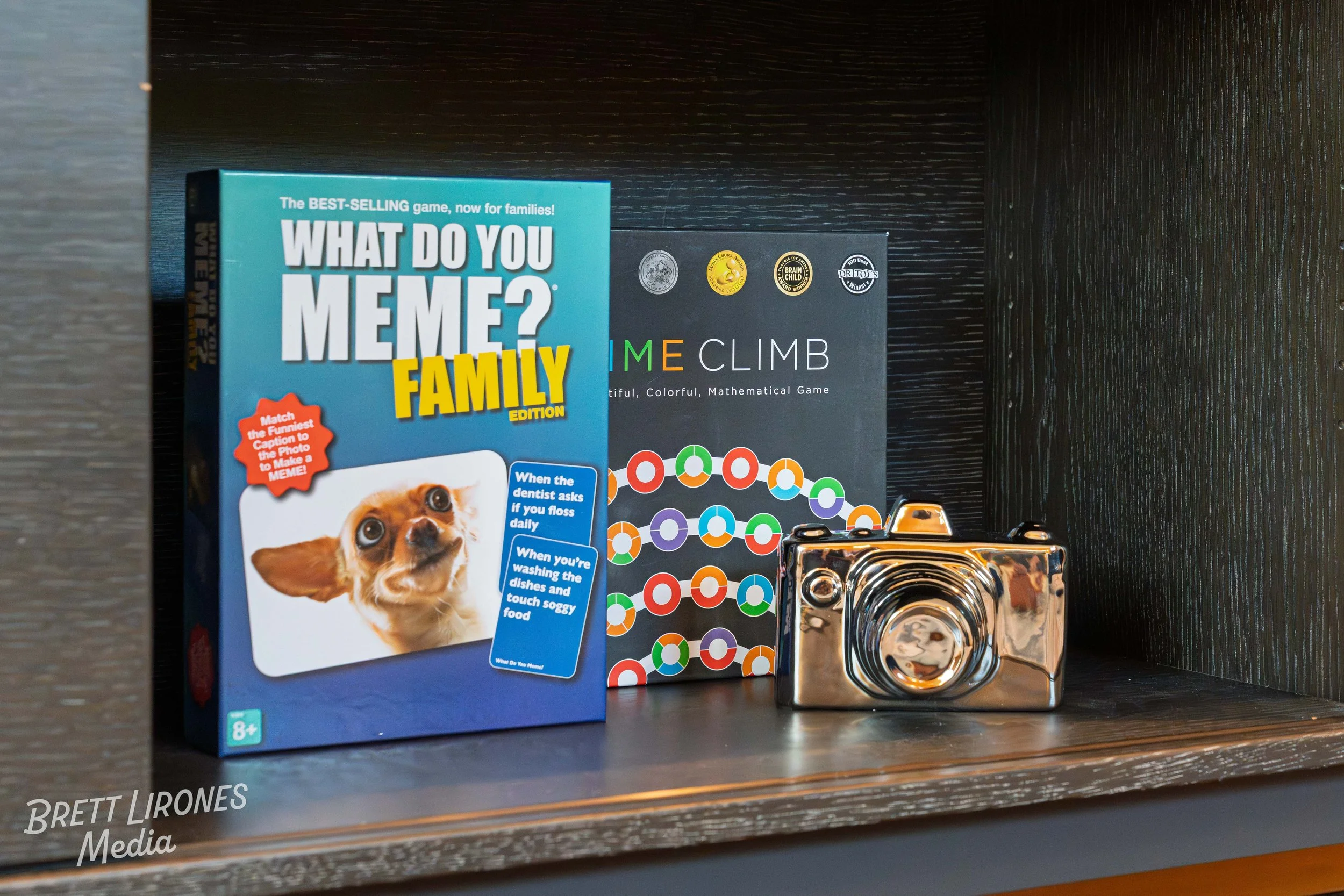 Shelf with three board games: 'What Do You Meme? Family Edition,' 'Game Climb,' and another with colorful circles, and a shiny metallic toy camera.