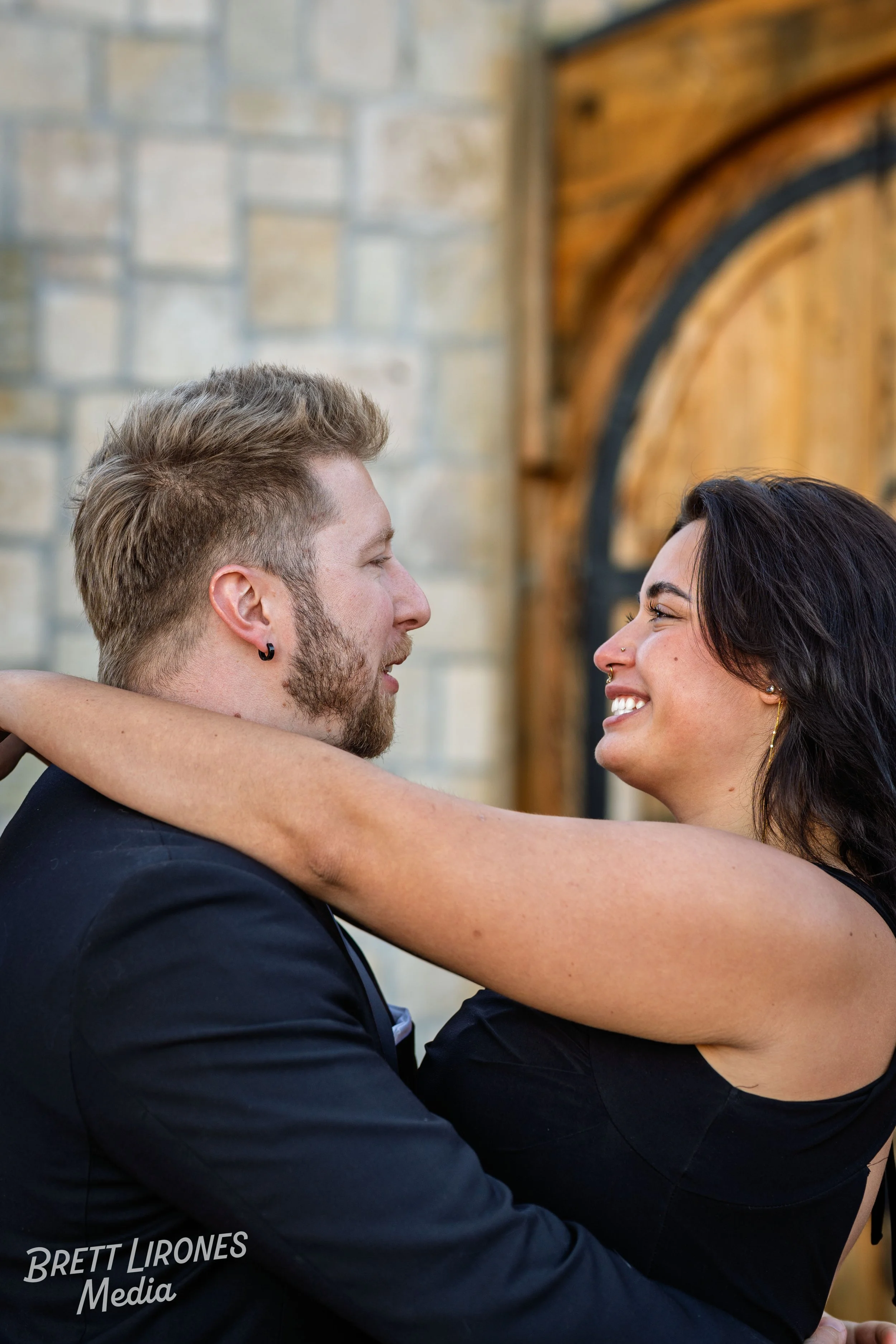 A couple embraces and looks at each other happily, with the woman smiling and the man looking lovingly. They are indoors with a rustic brick and wood background.