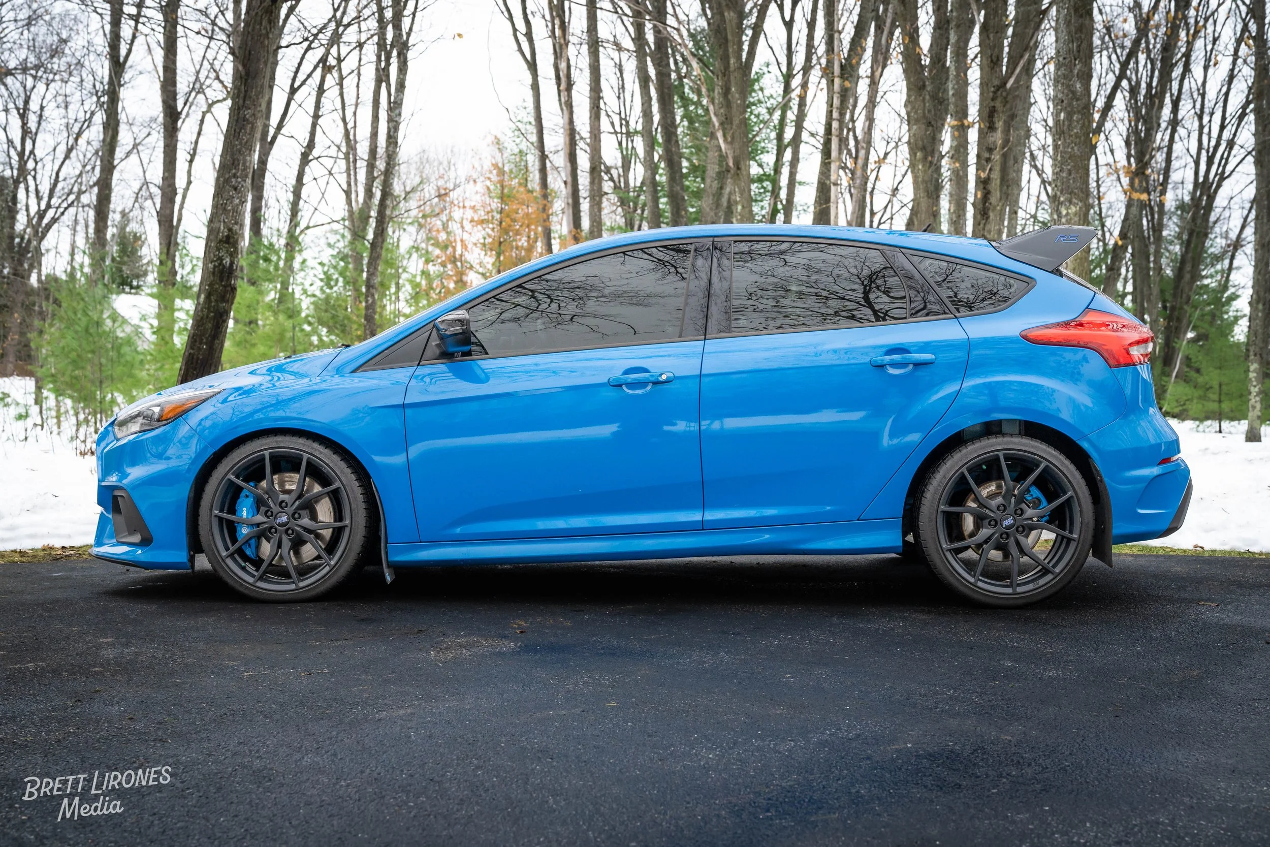 Blue hatchback car parked on a paved surface in front of a wooded area with snow, trees, and some green foliage.