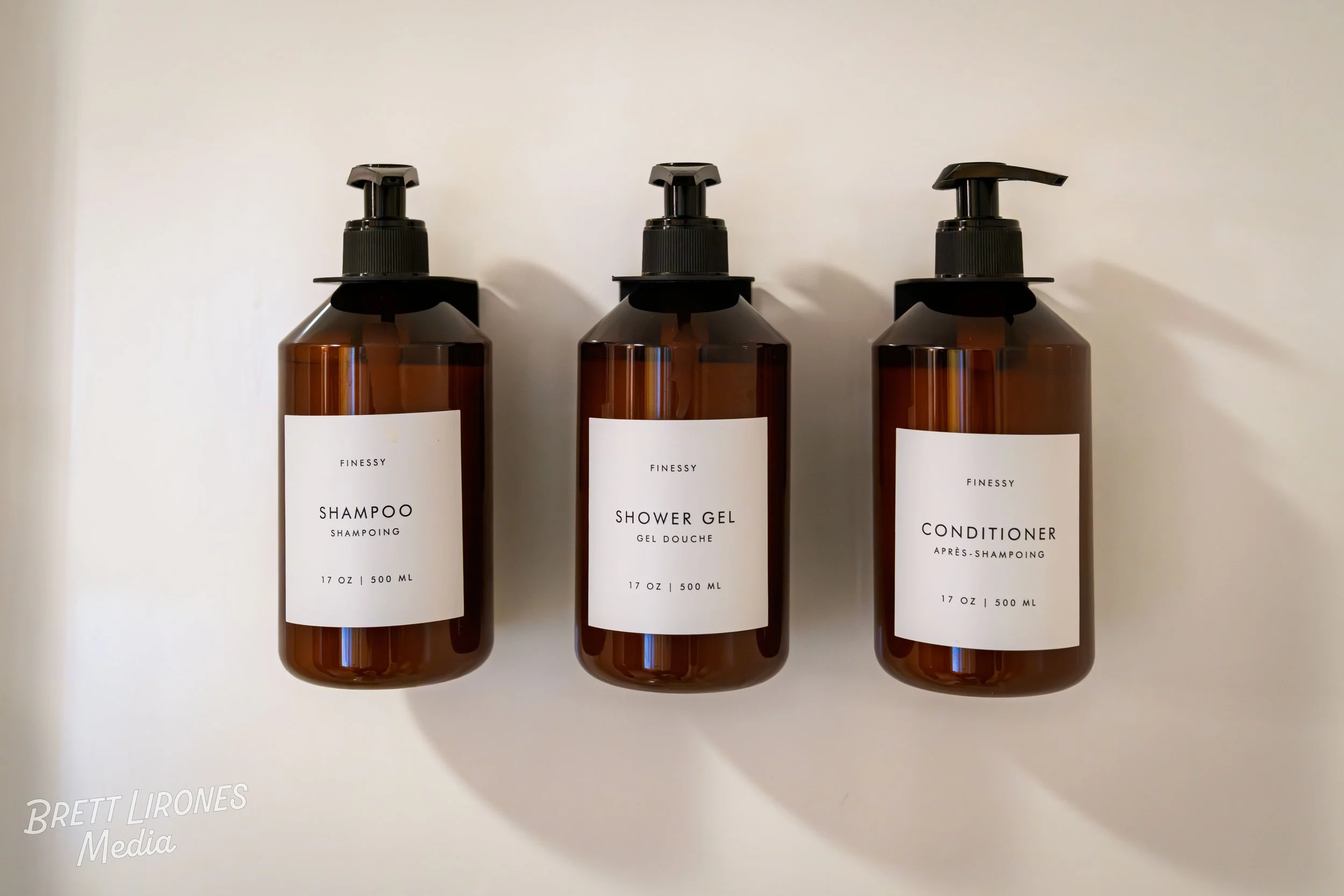 Three brown plastic bottles with black pump tops on a white surface. The bottles are labeled 'Finesse,' containing shampoo, shower gel, and conditioner, each 17 ounces or 500 milliliters.