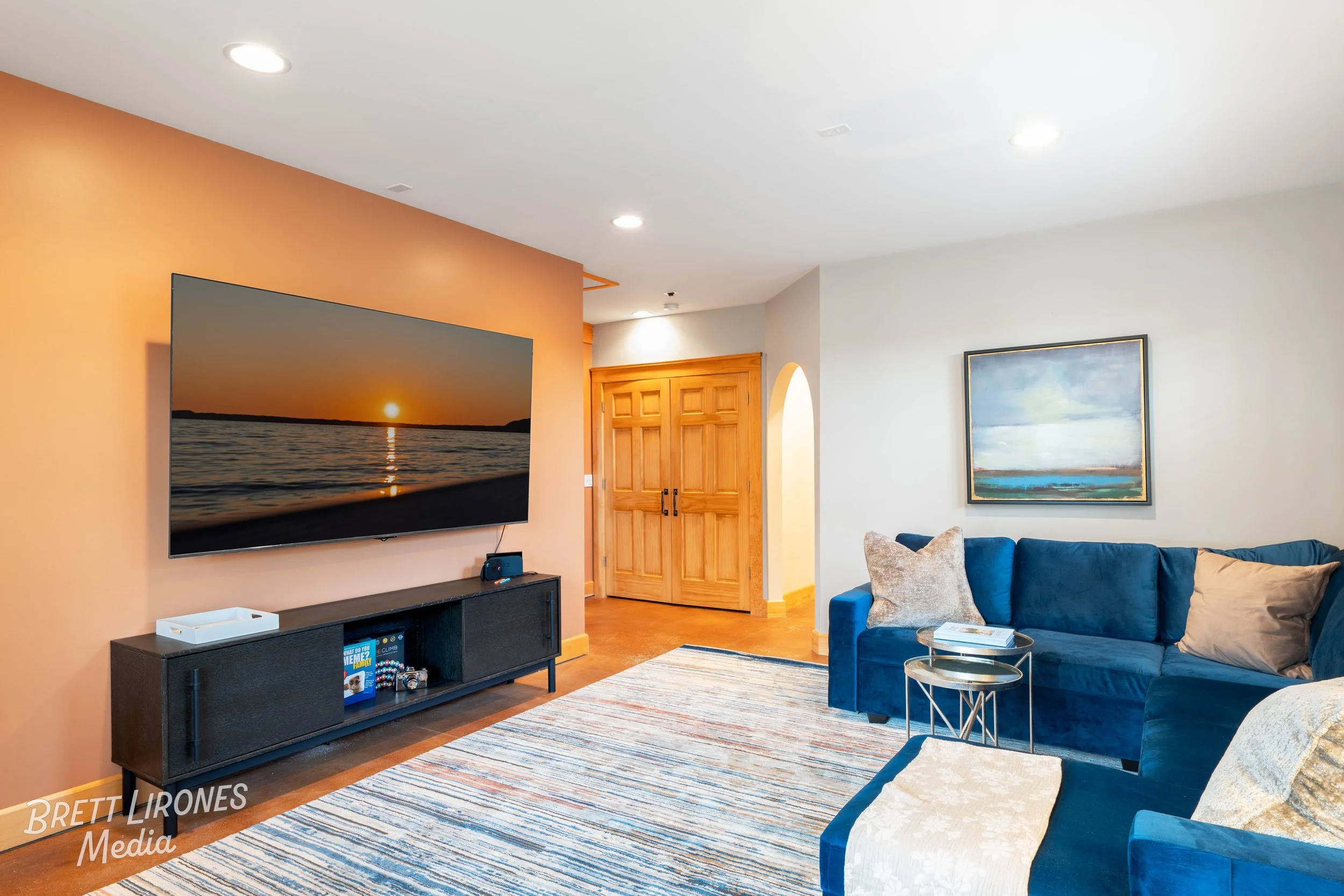 Living room with a large wall-mounted TV showing a sunset over water, blue sofa with beige and decorative pillows, colorful striped rug, wooden door, and framed landscape painting on white wall