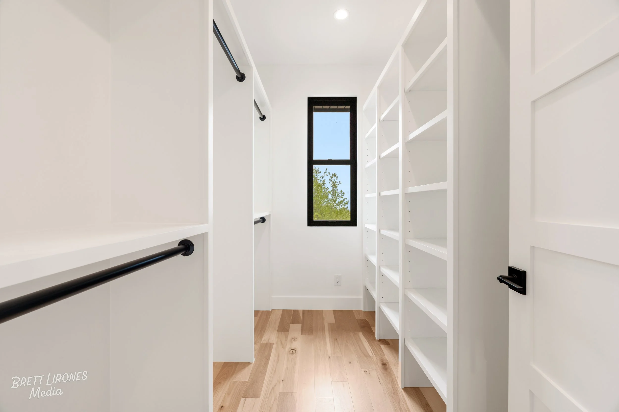 Empty walk-in closet with white shelves and black rods, natural light coming through a narrow window, light wooden flooring, and a white door.