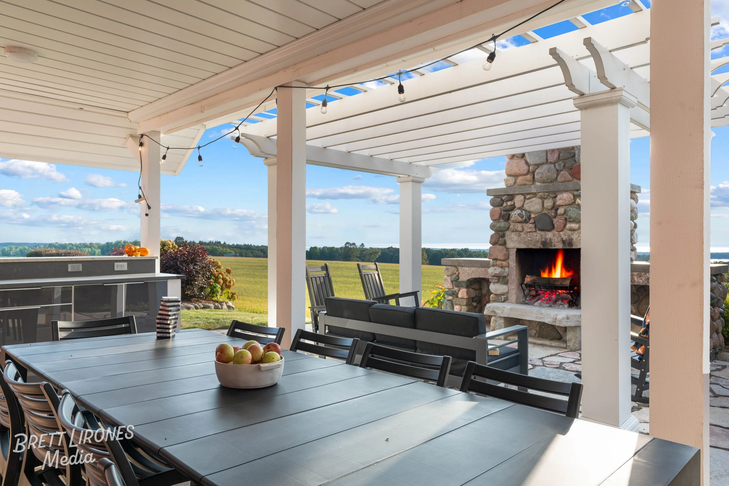 Covered outdoor patio with a large dining table, a stone fireplace with fire burning, and chairs, overlooking open green fields and blue sky.
