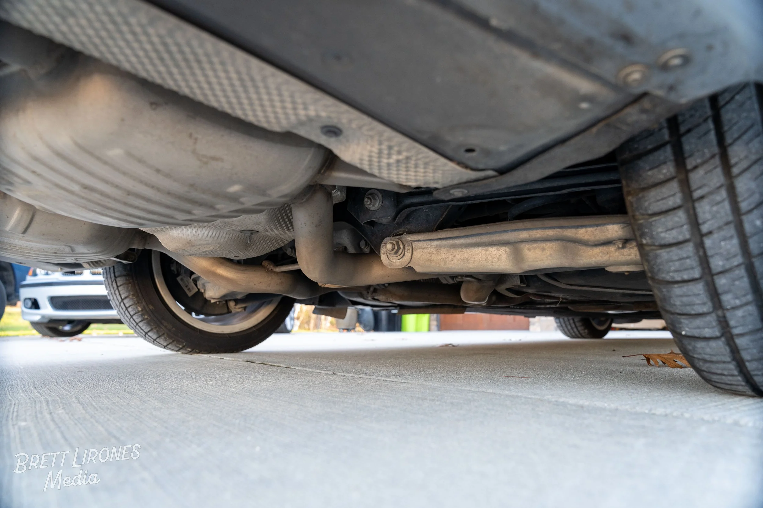 Underneath view of a car showing the exhaust system, suspension, and tires parked on a concrete driveway.