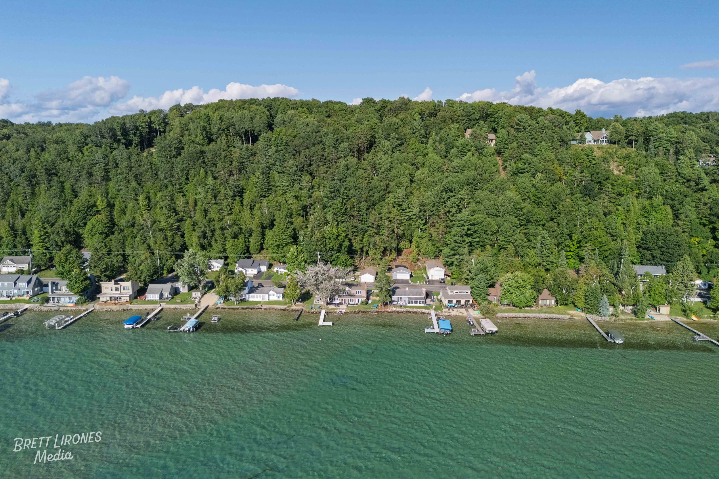 Aerial view of a lakeside community with boats at docks, houses, and a forested hill in the background under a partly cloudy sky.