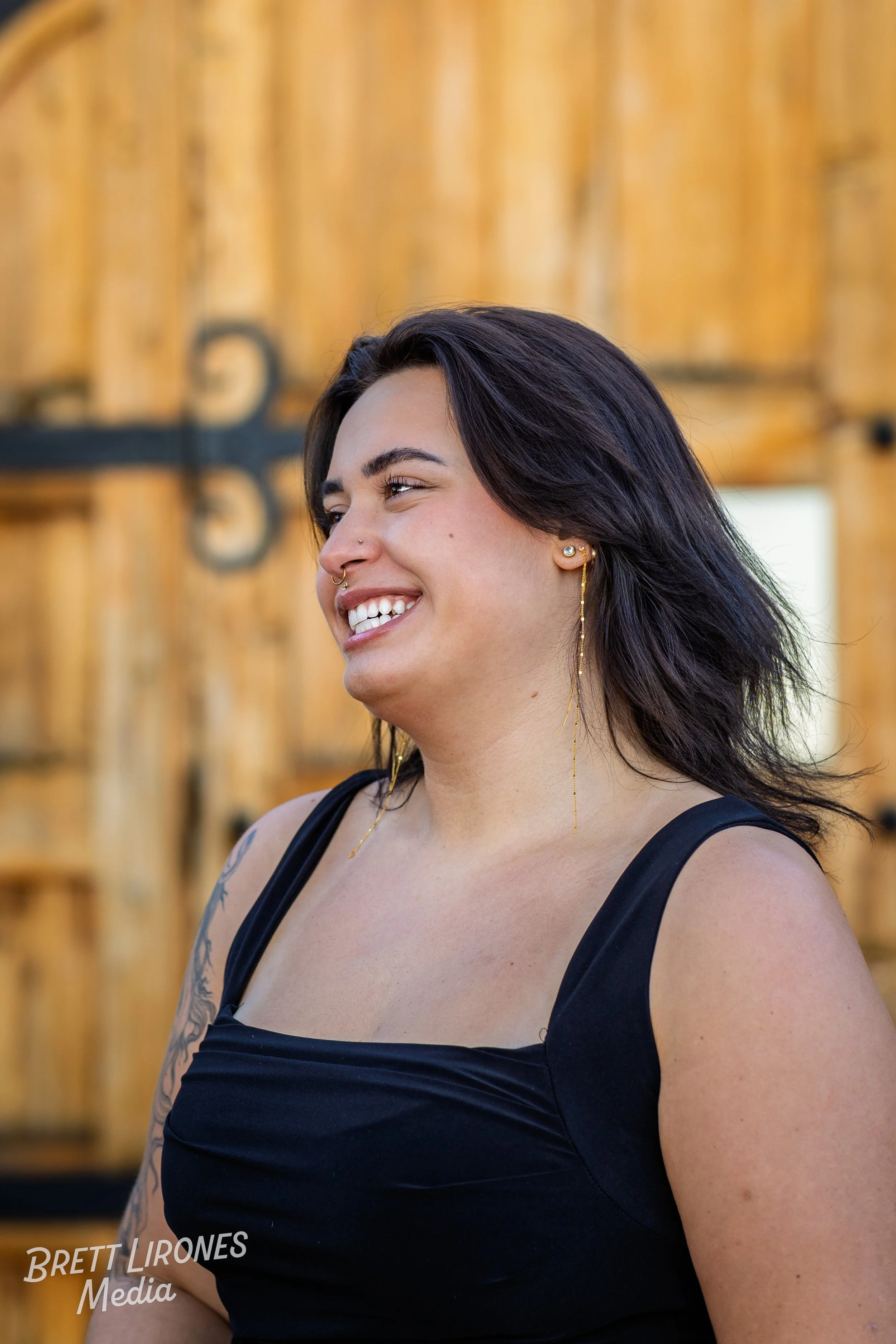 A woman with dark hair and tattoos smiling outdoors with a wooden background.