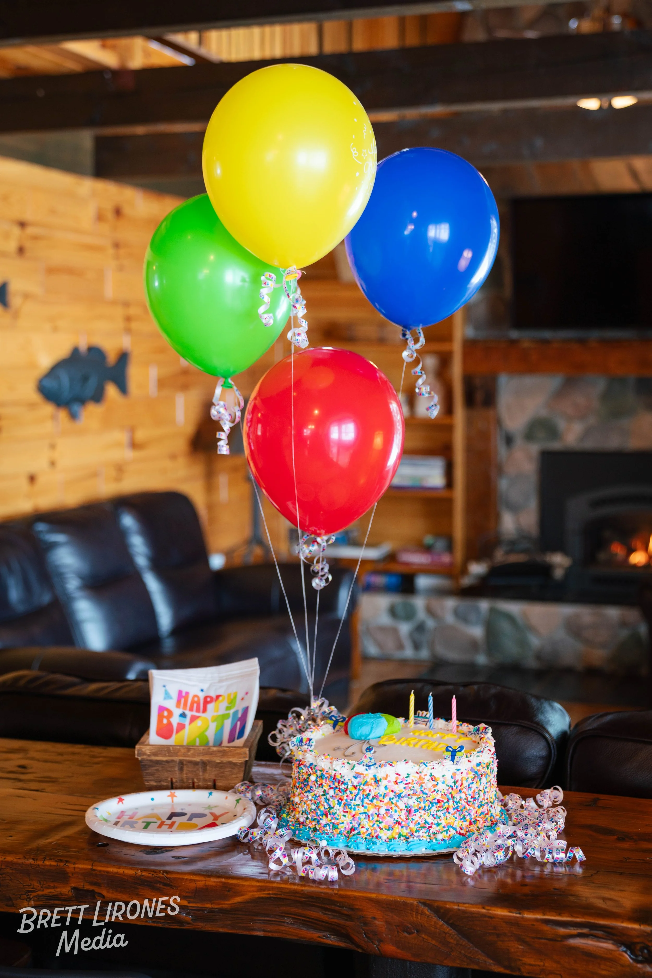 Colorful birthday balloons over a birthday cake with rainbow sprinkles on a wooden table, with a 'Happy Birthday' sign and birthday plate nearby, in a cozy room with wood panel walls and a stone fireplace.