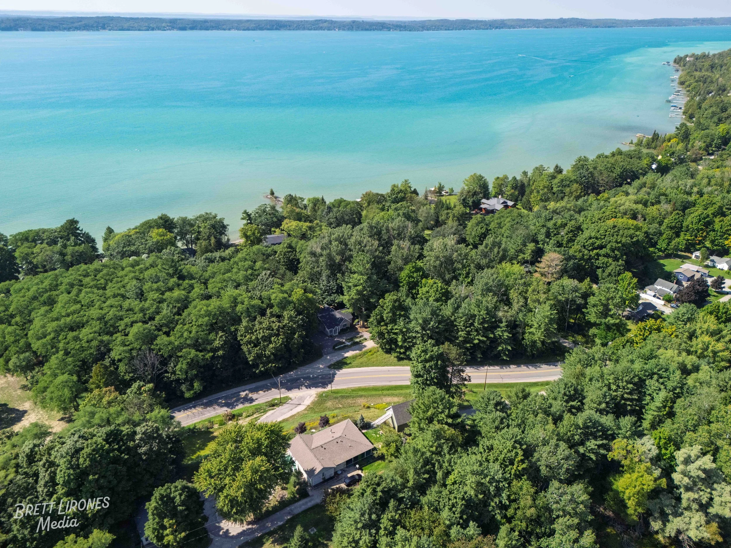 Aerial view of a coastal neighborhood with lush green trees, houses, and a winding road near a large, blue lake or sea.