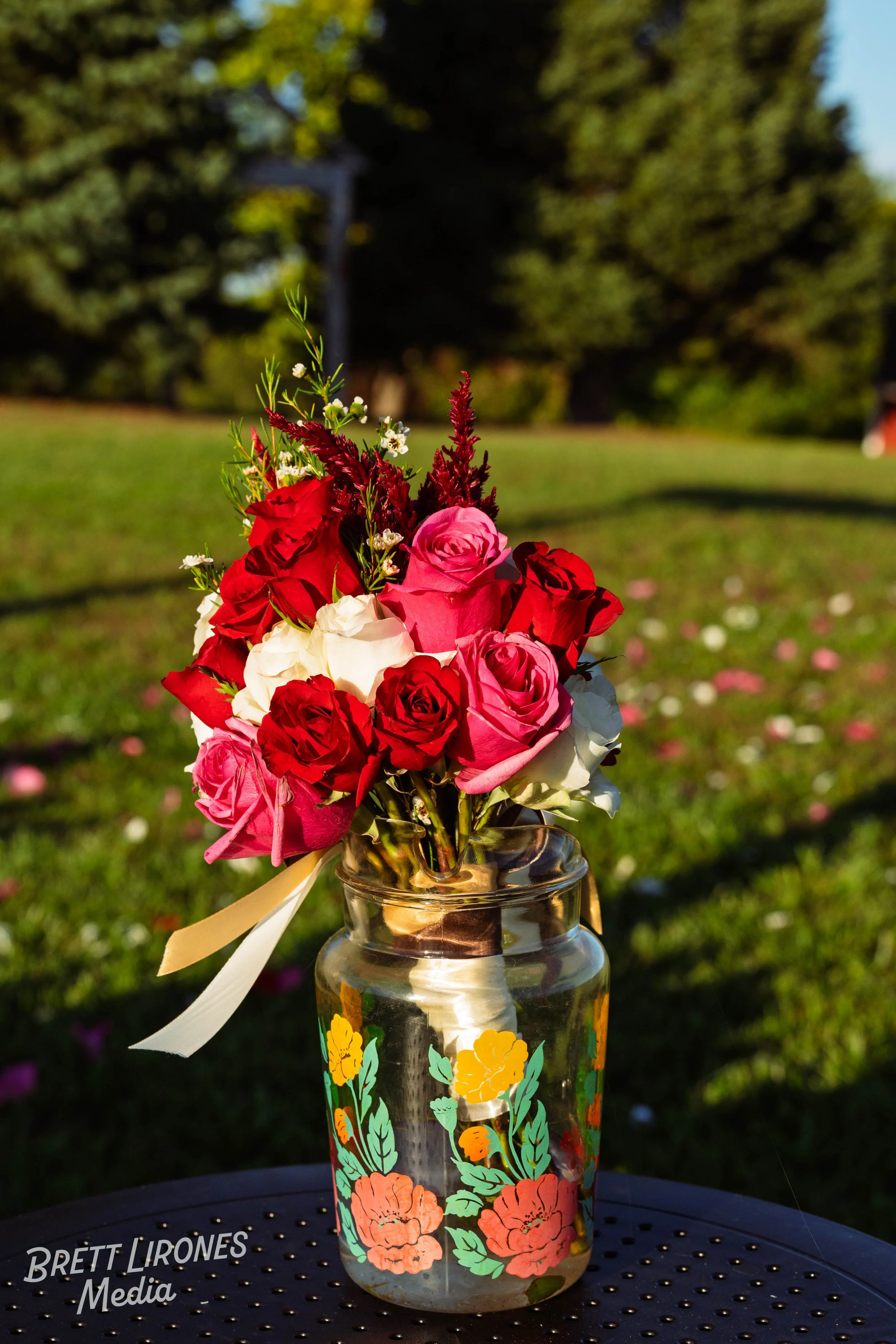 A bouquet of red, pink, and white roses in a decorated glass jar, outdoors on a sunny day.