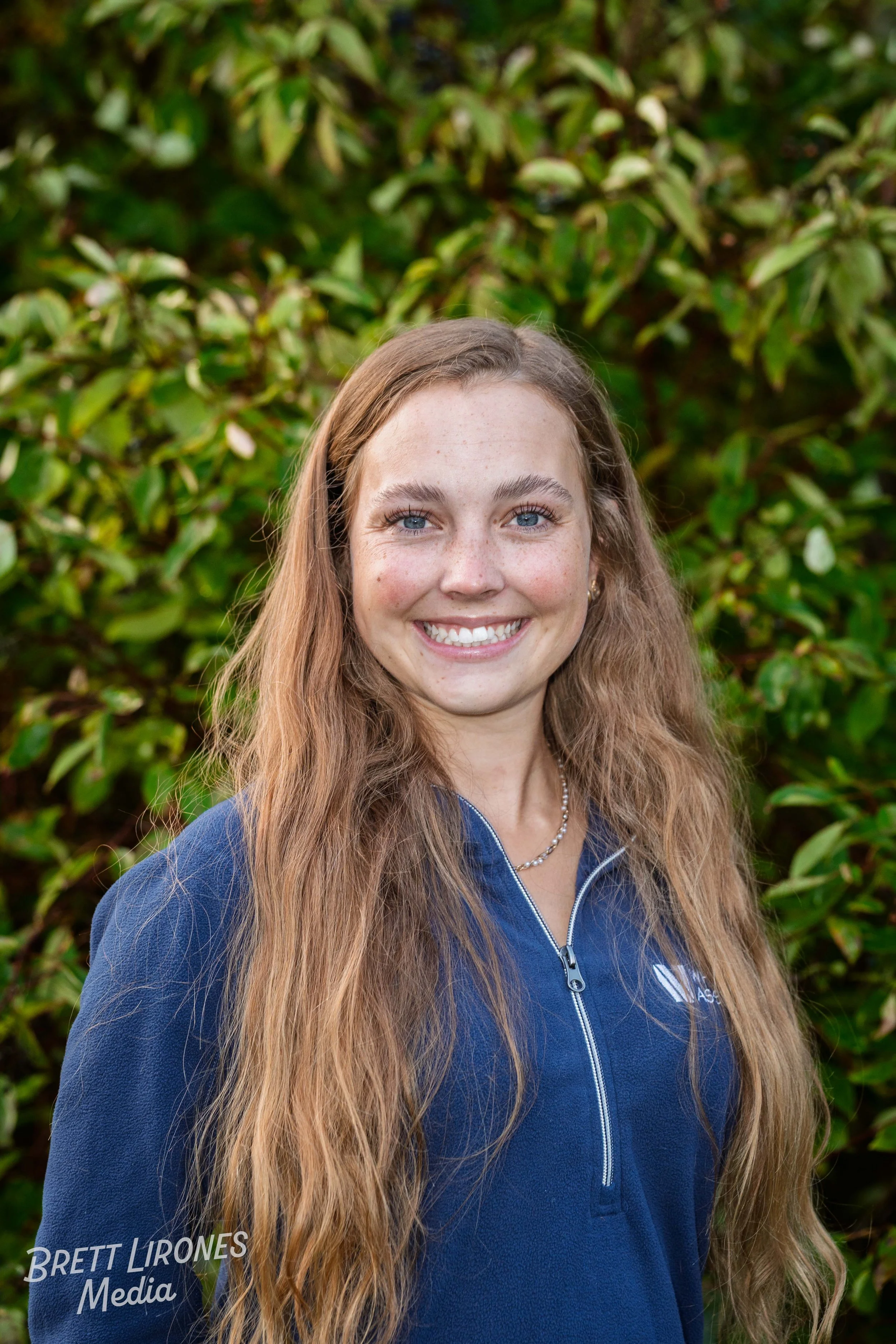 A young woman with long, wavy light brown hair, blue eyes, and freckles, smiling in front of green leafy bushes. She is wearing a navy blue zip-up jacket with a logo and a pearl necklace.