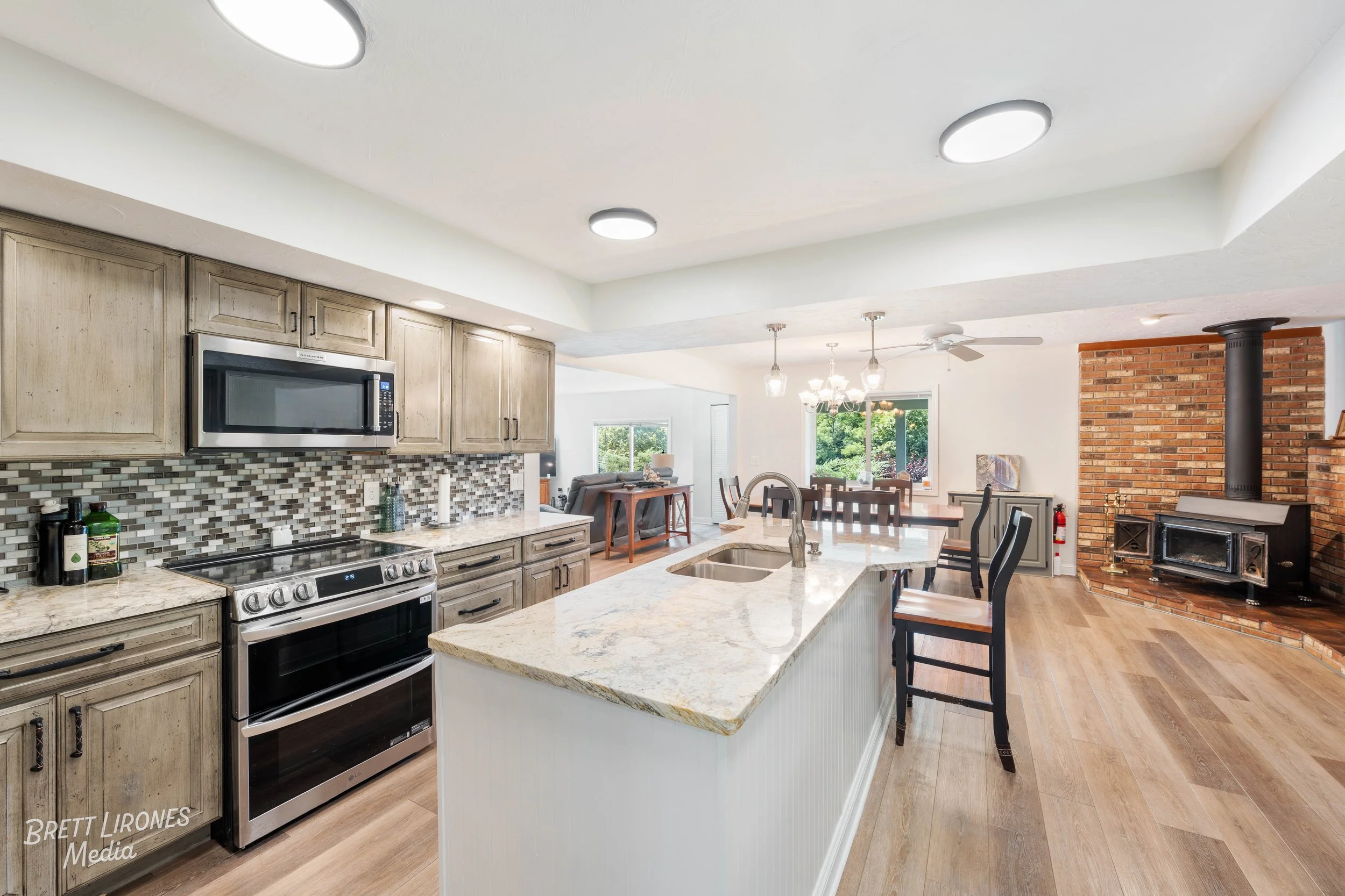 Open-concept kitchen with beige cabinets, granite countertops, mosaic backsplash, and stainless steel stove and microwave, flowing into dining and living areas with wooden floors and brick fireplace.