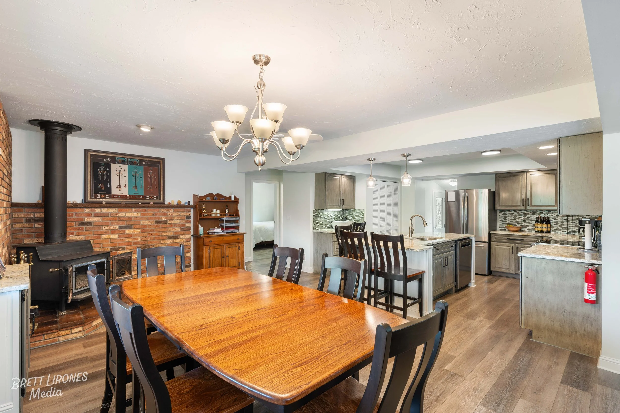 Open-concept dining and kitchen area with a wooden dining table, black chairs, a brick fireplace, and gray cabinetry in the kitchen.