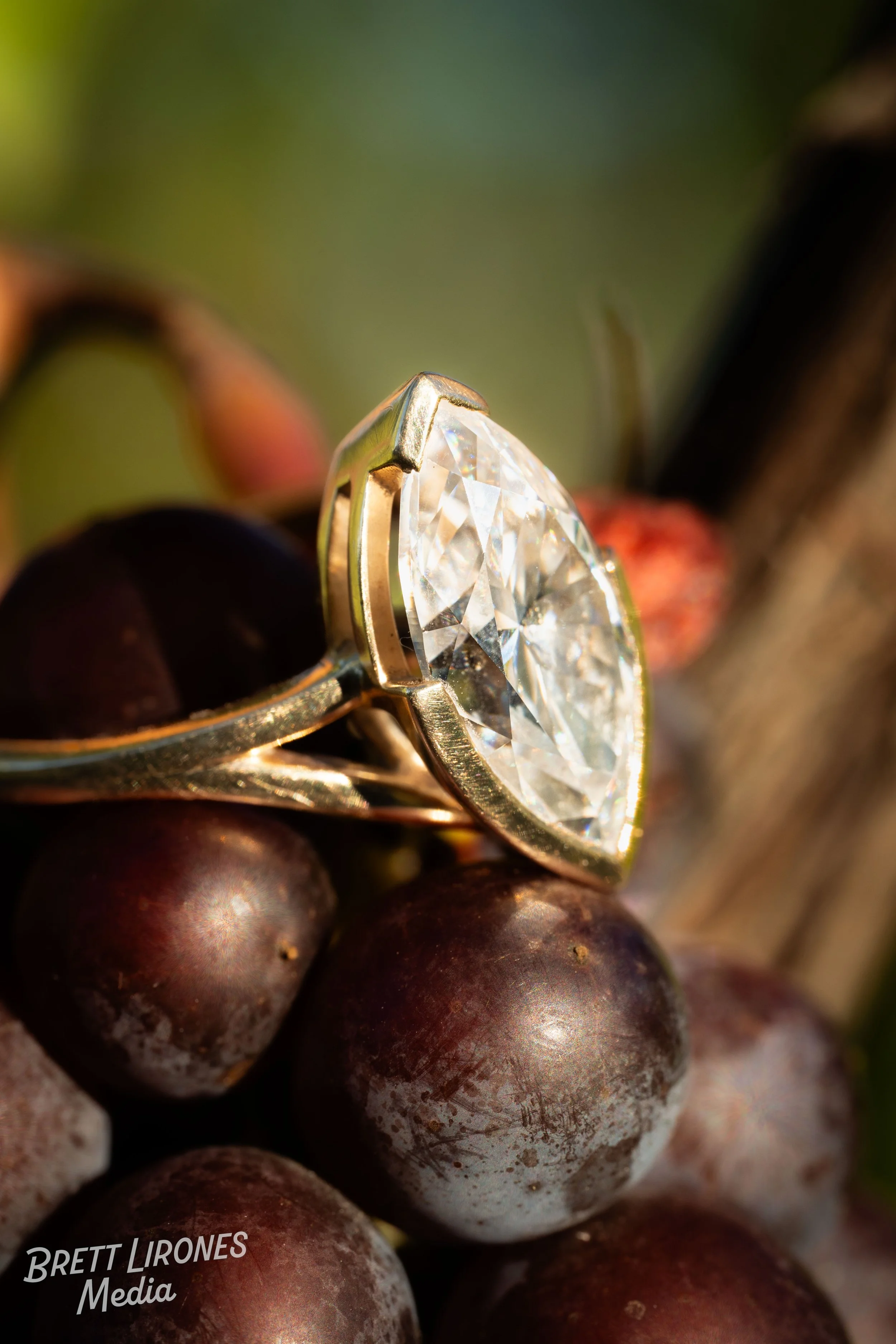 Close-up of a diamond engagement ring with a gold band resting on dark purple grapes.