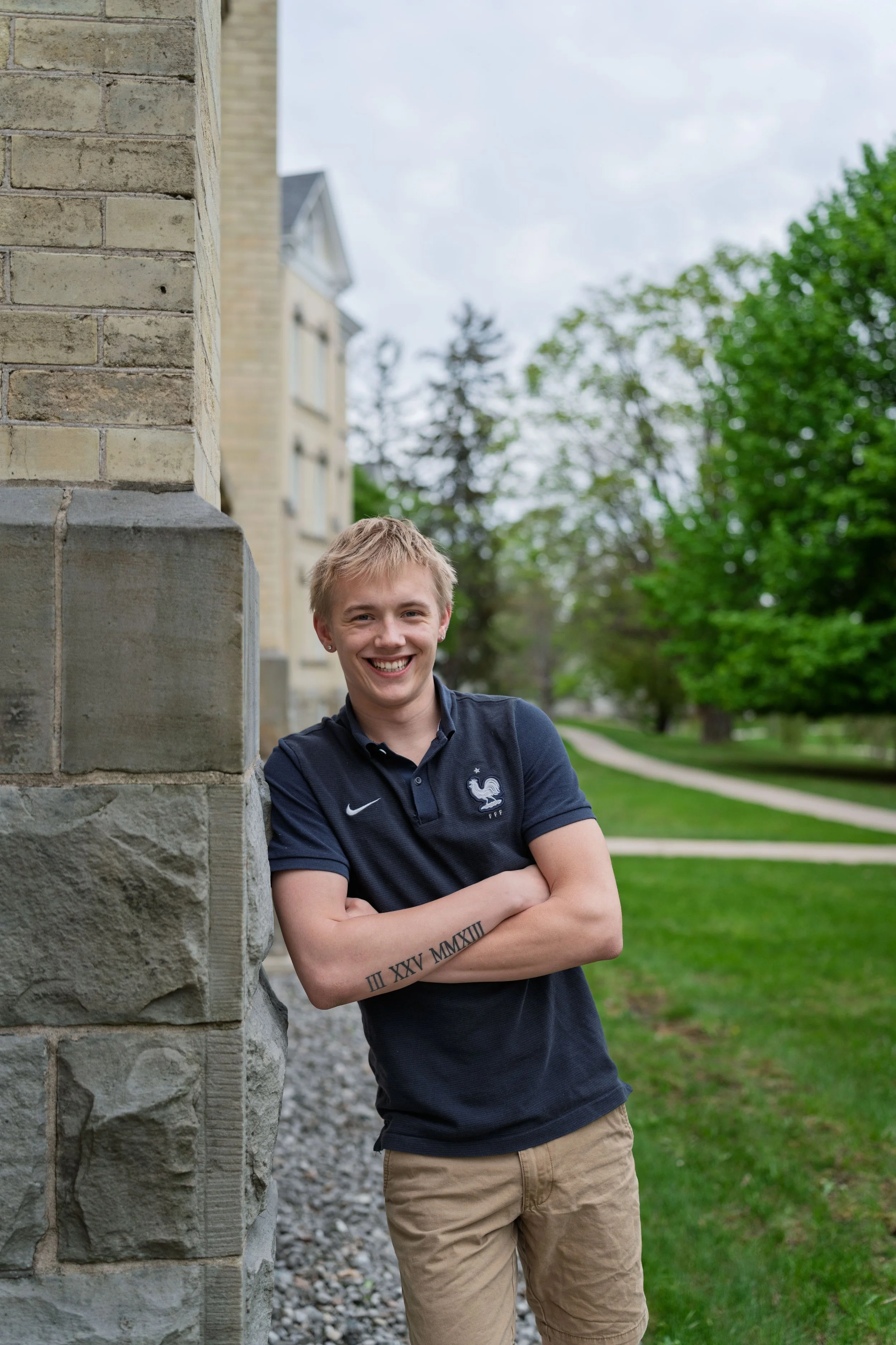 A young man with blond hair smiling, standing outdoors with arms crossed, leaning against a stone building, with trees and a grassy area in the background.
