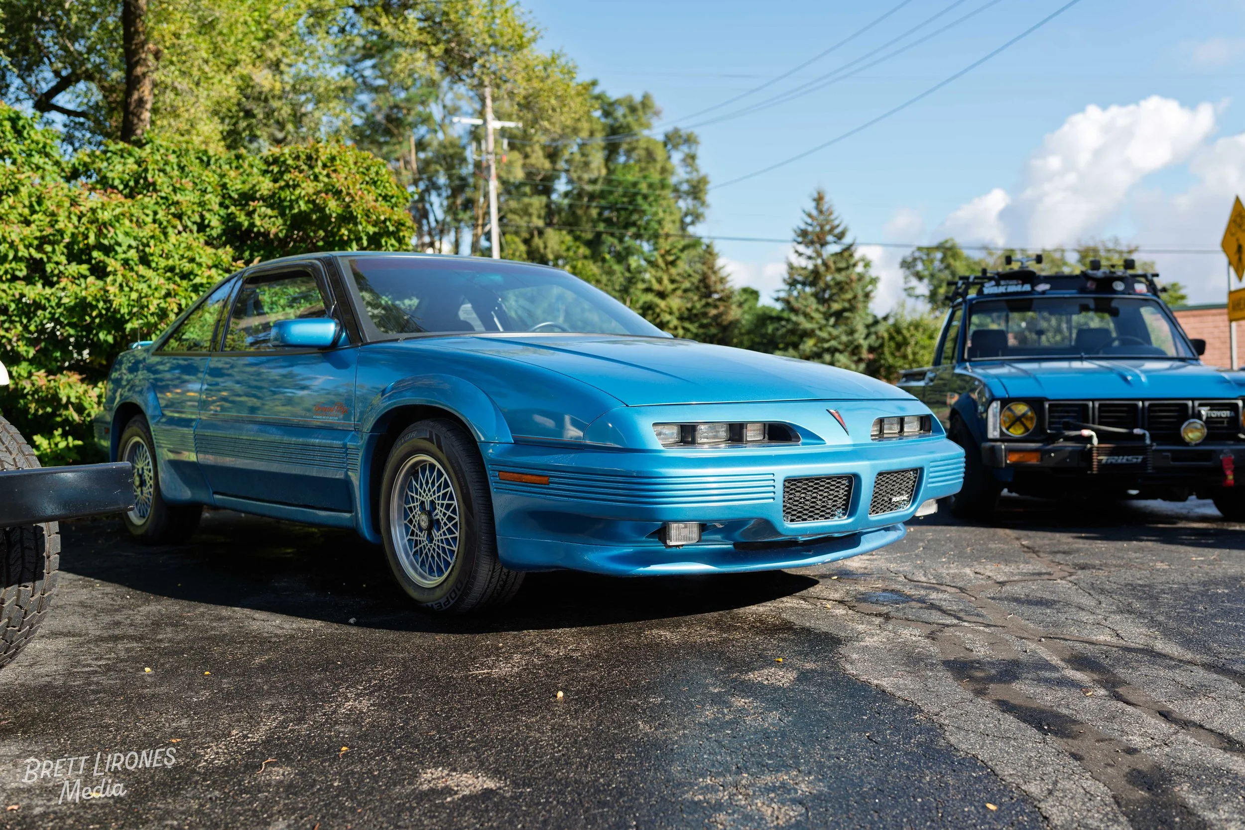 A blue vintage sports car parked on a brick-paved lot with green trees in the background, next to a grey off-road vehicle.