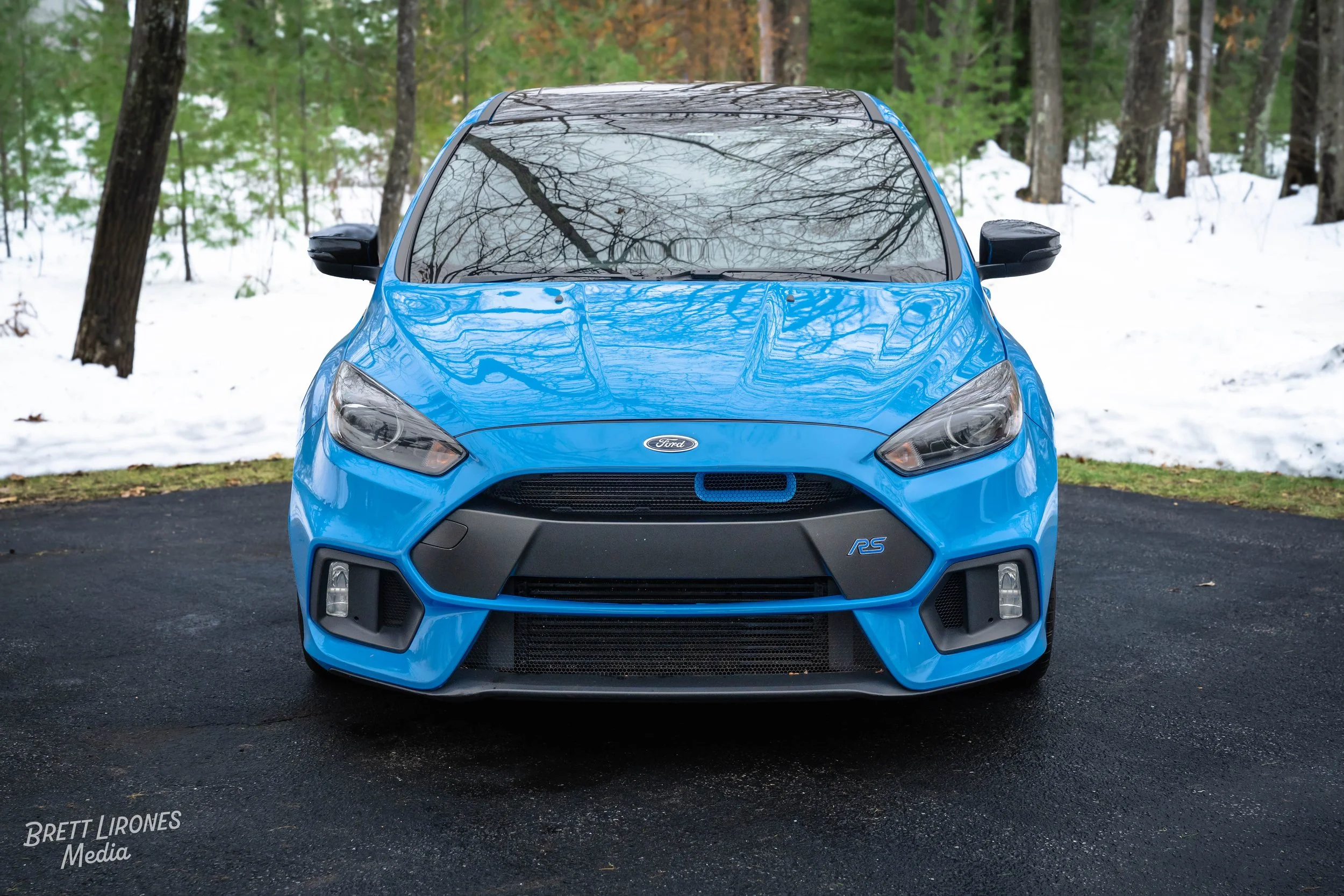 Front view of a blue Ford Focus RS parked on a paved driveway with snow and trees in the background.