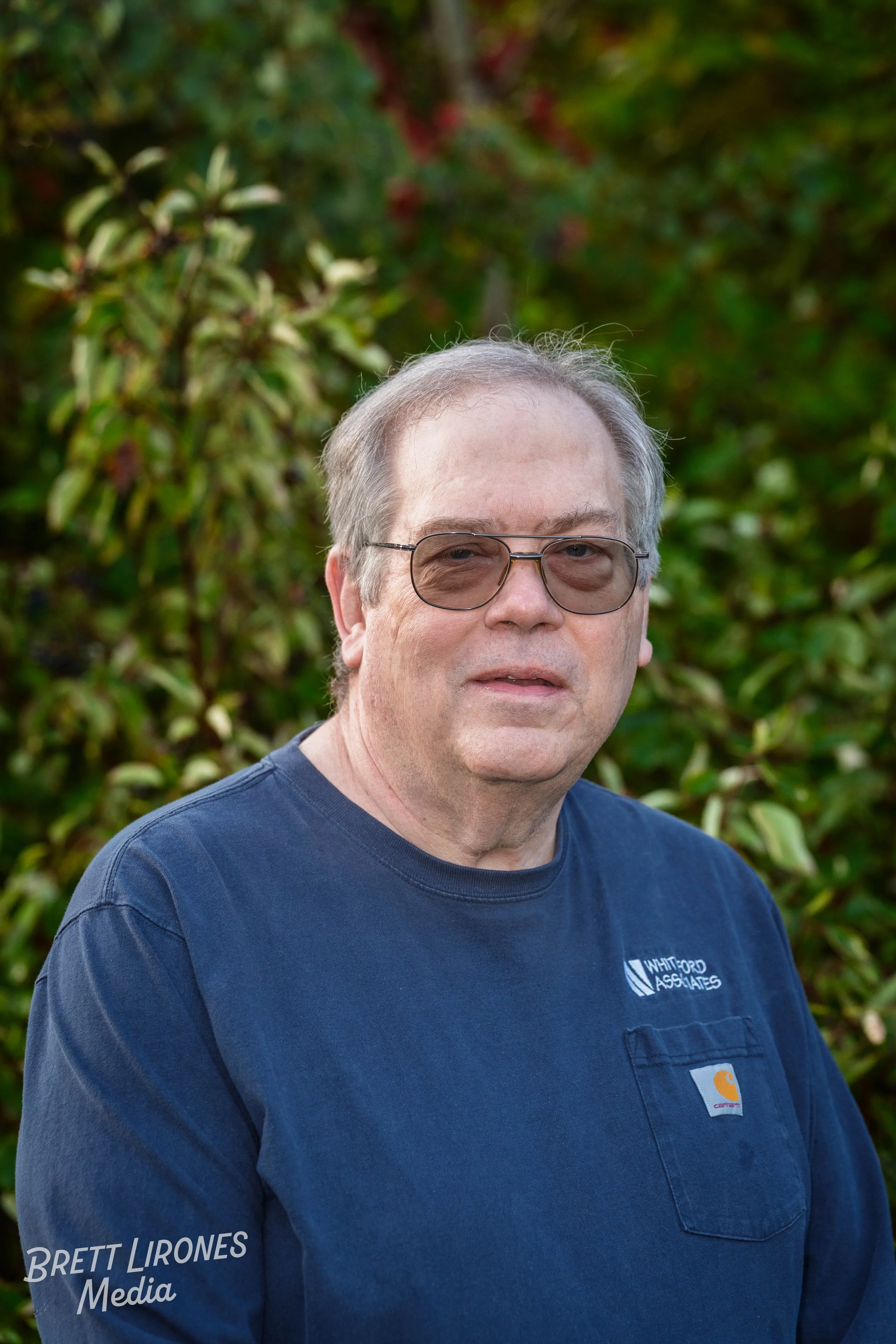 A middle-aged man with gray hair, glasses, wearing a navy blue shirt with logos, standing outdoors with greenery in the background.