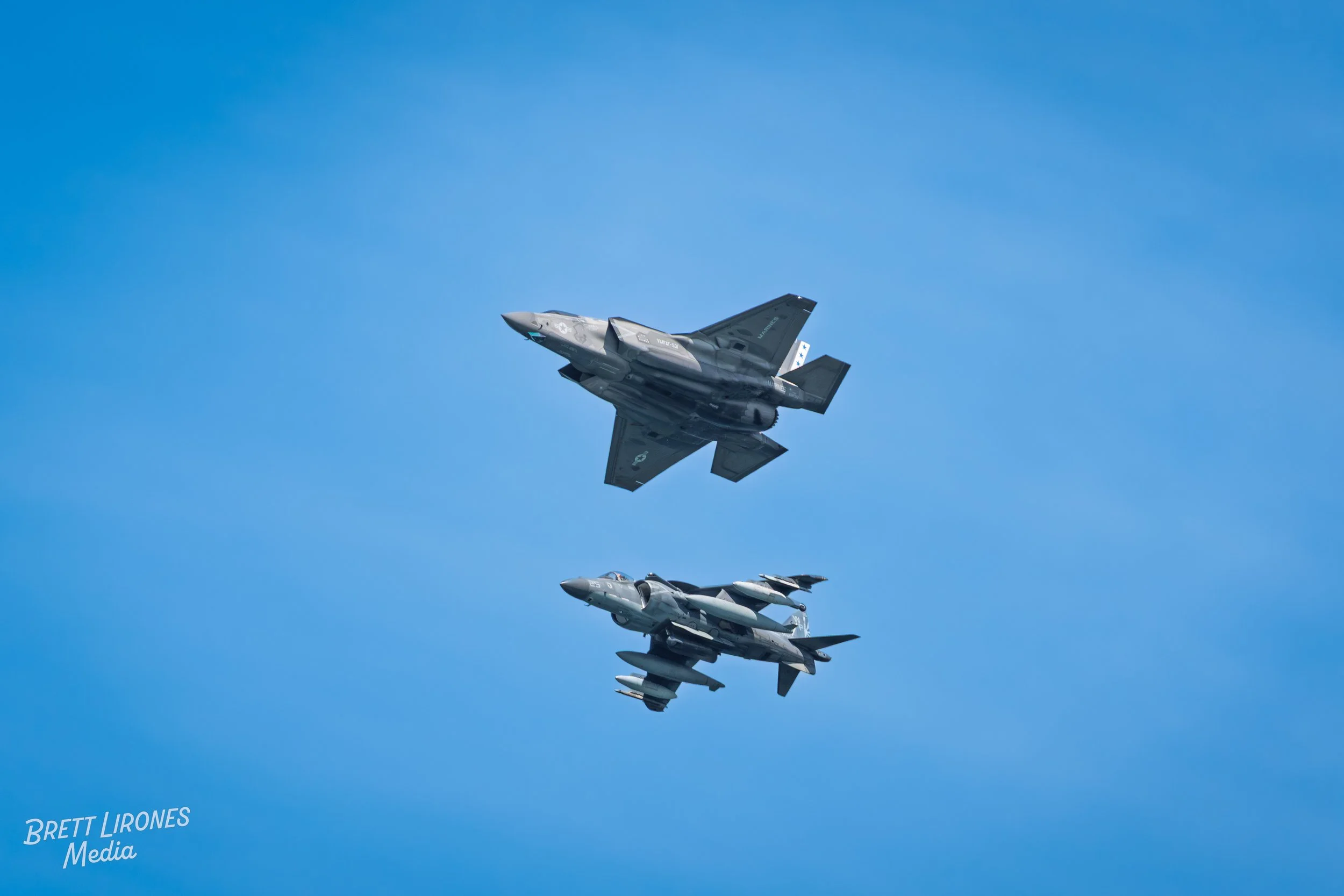 Two fighter jets flying in clear blue sky, one above the other, with the words 'BRETT LIRONES Media' in the bottom left corner.
