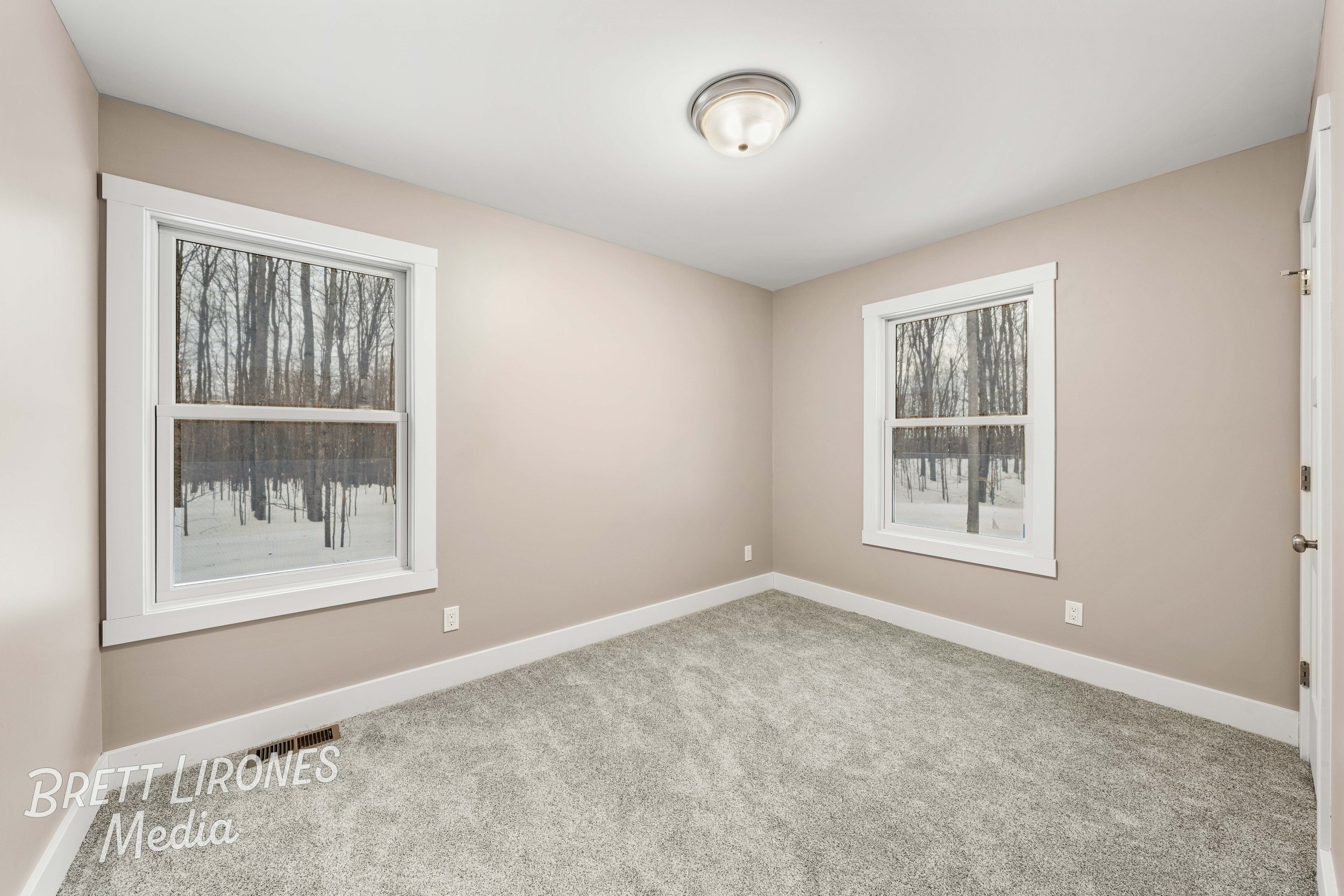 Empty room with beige walls, carpeted floor, two large windows showing a snowy wooded landscape, white window frames and baseboards, ceiling light fixture.