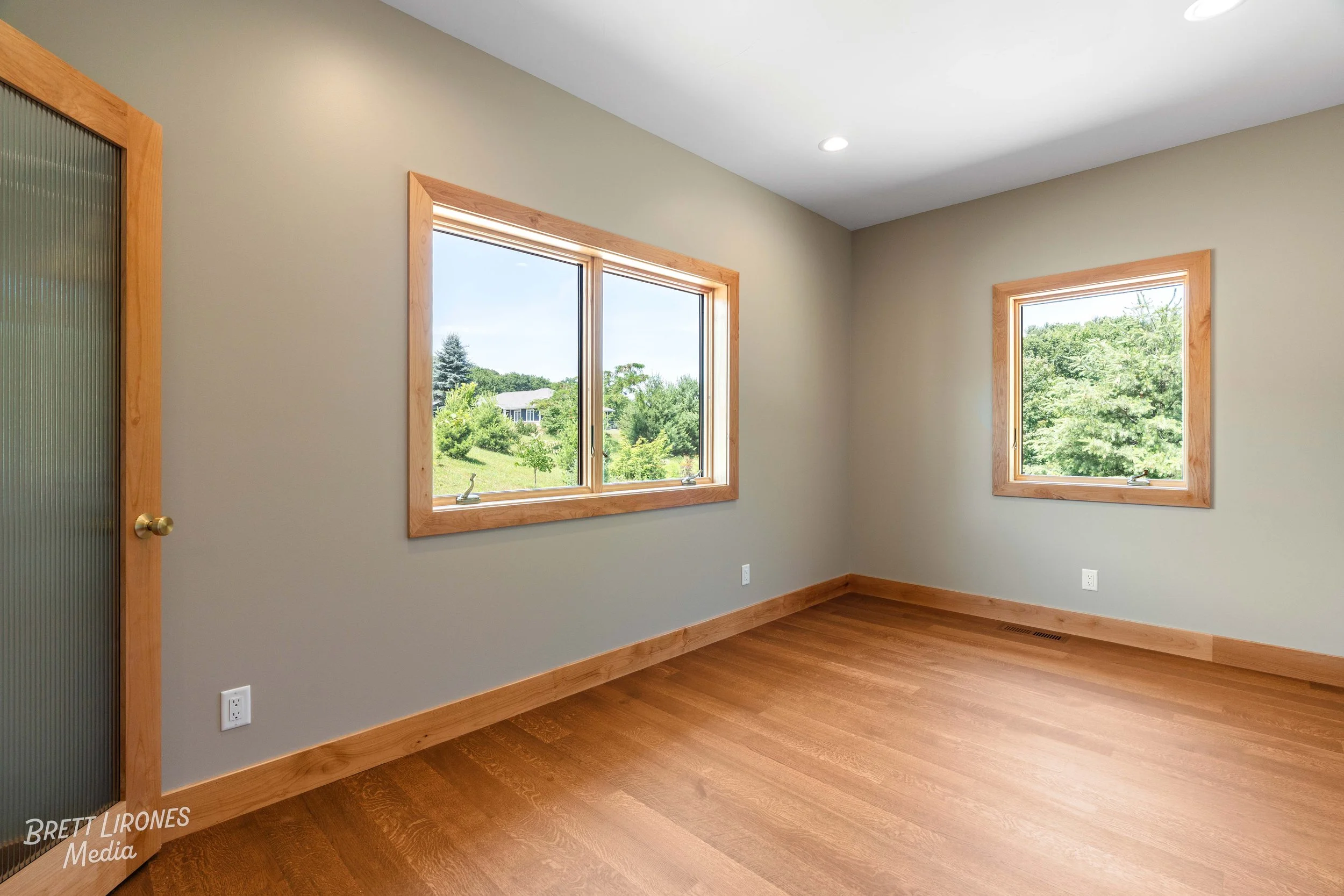 Empty room with two large windows showing green trees and a yard outside, beige walls, wood trim and baseboards, hardwood flooring, and a door with textured glass and a brass doorknob.