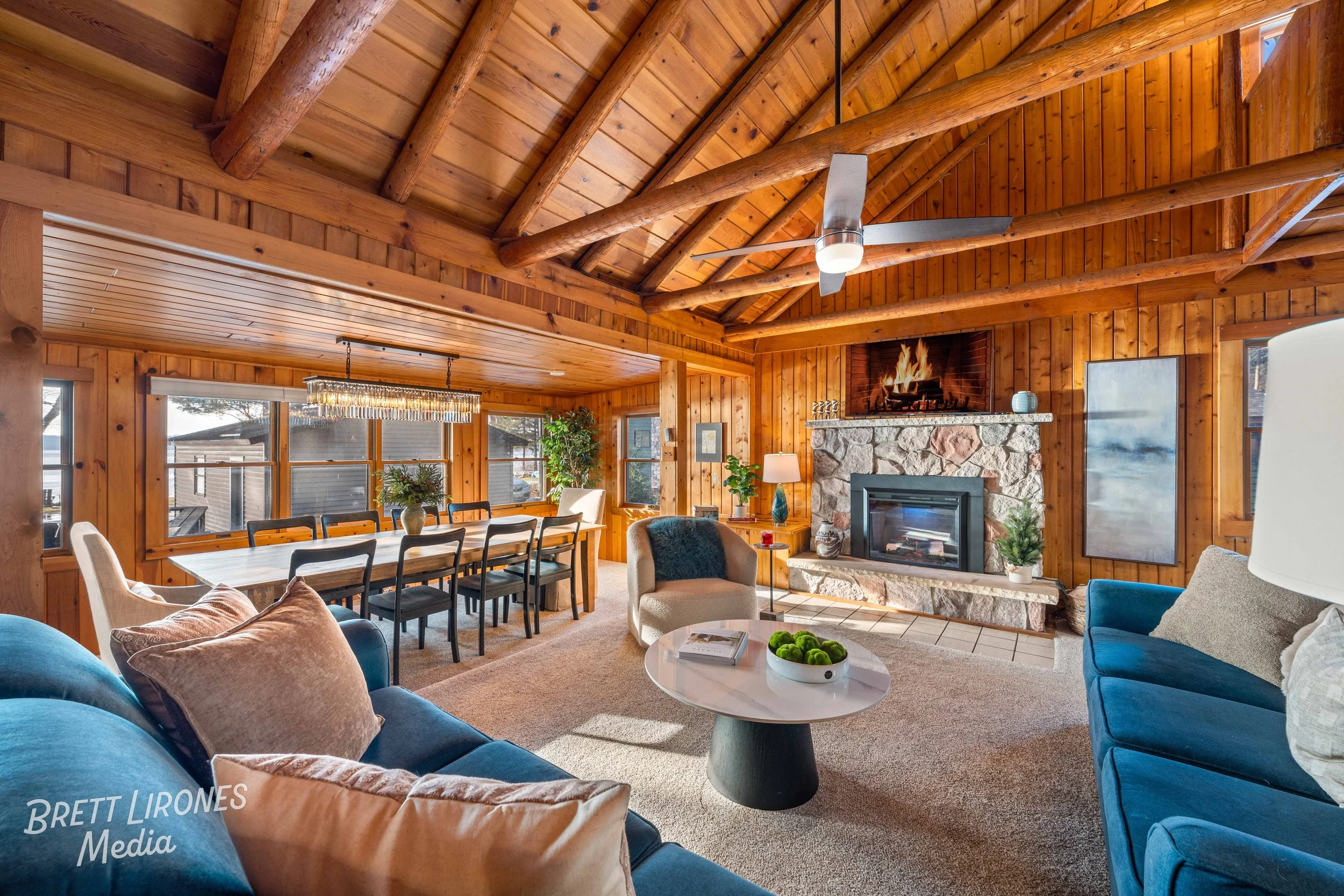 Interior view of a cozy living room with wooden walls and ceiling, a stone fireplace with a fire, a blue sofa, beige armchair, and a white coffee table with a bowl of green apples, along with a dining area featuring a long table and chairs.