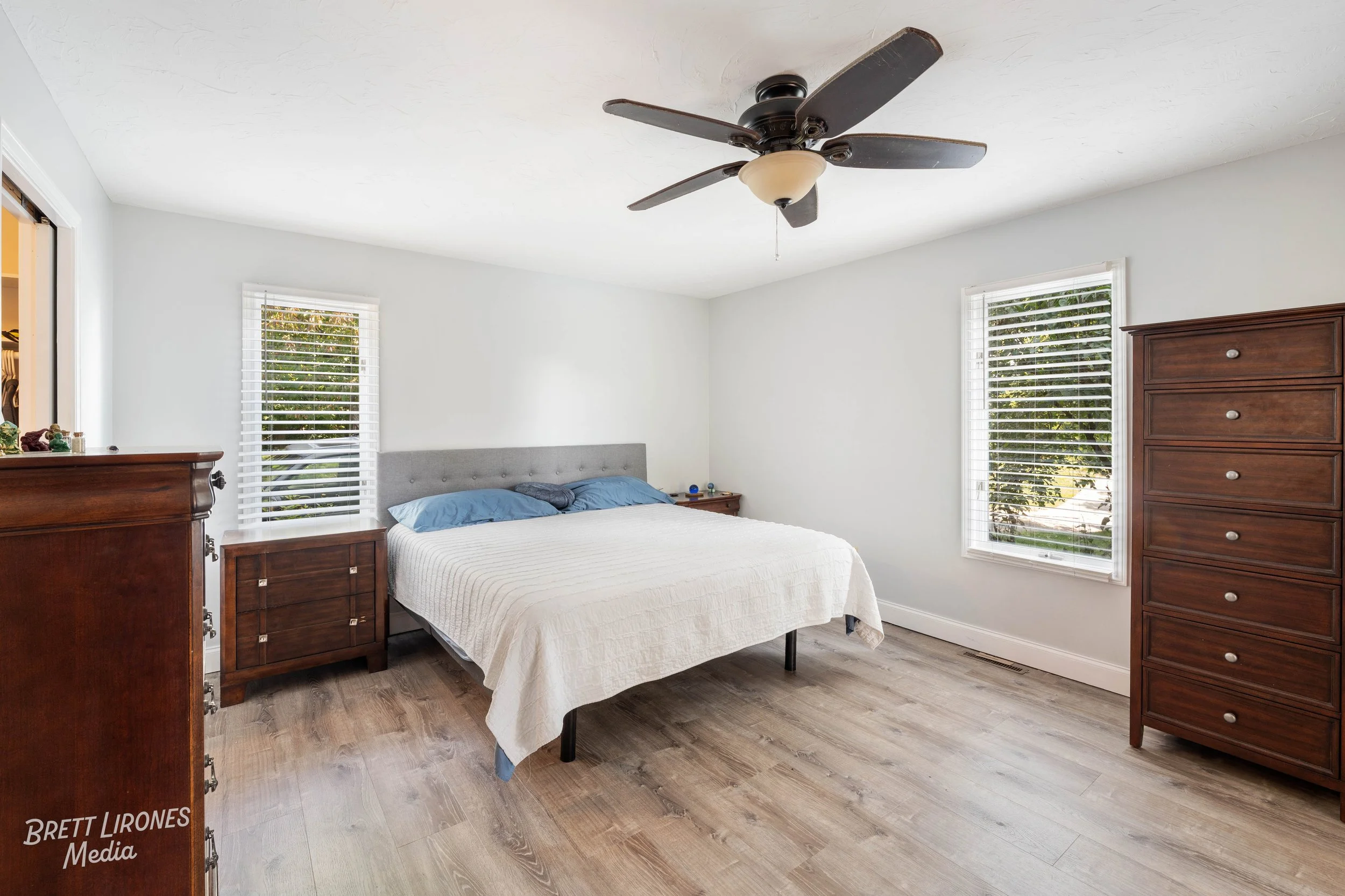 A bedroom with a bed, wooden dressers, and two windows with white blinds, ceiling fan, and wooden flooring.