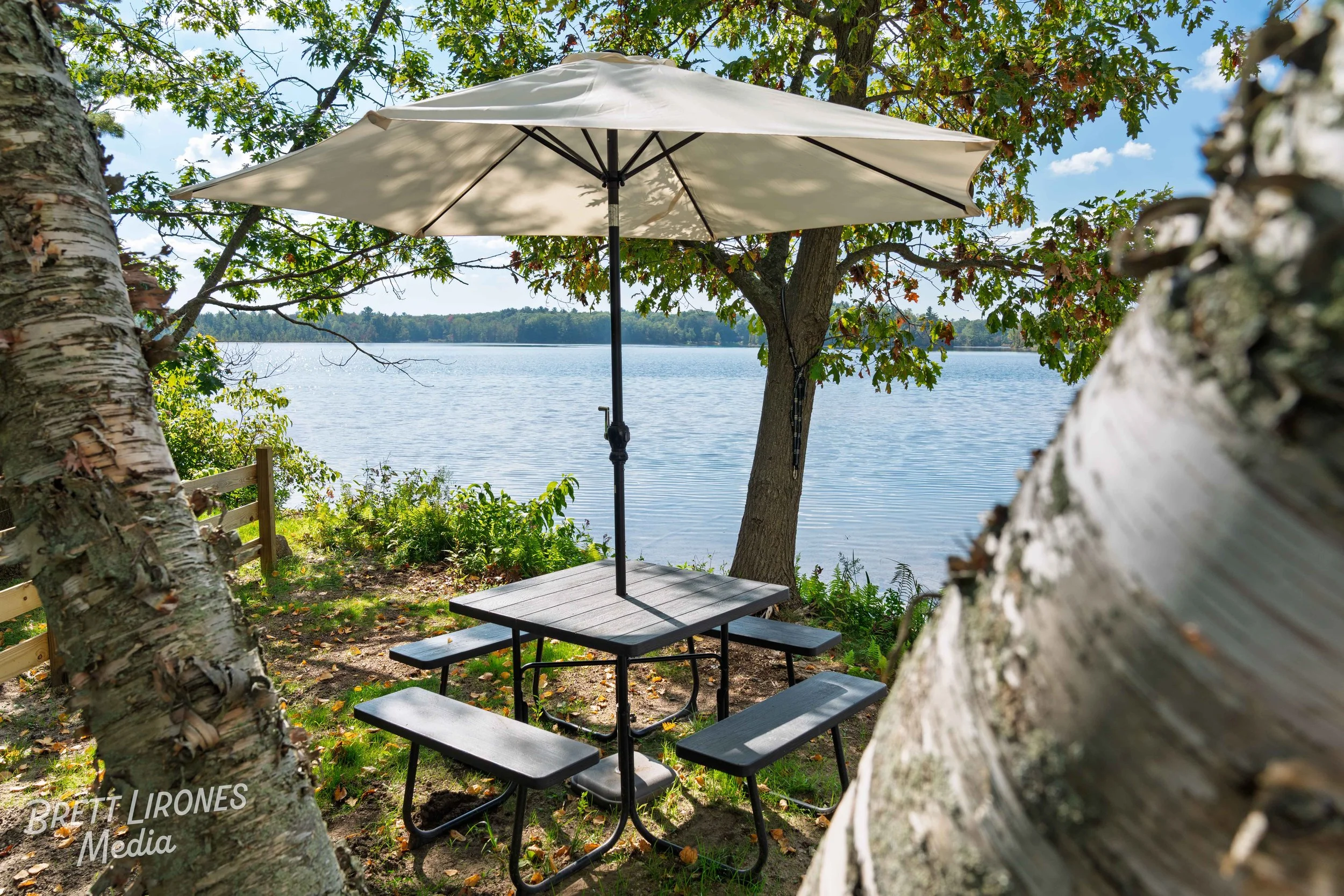 A picnic table with an umbrella near a lake, surrounded by trees with green leaves.