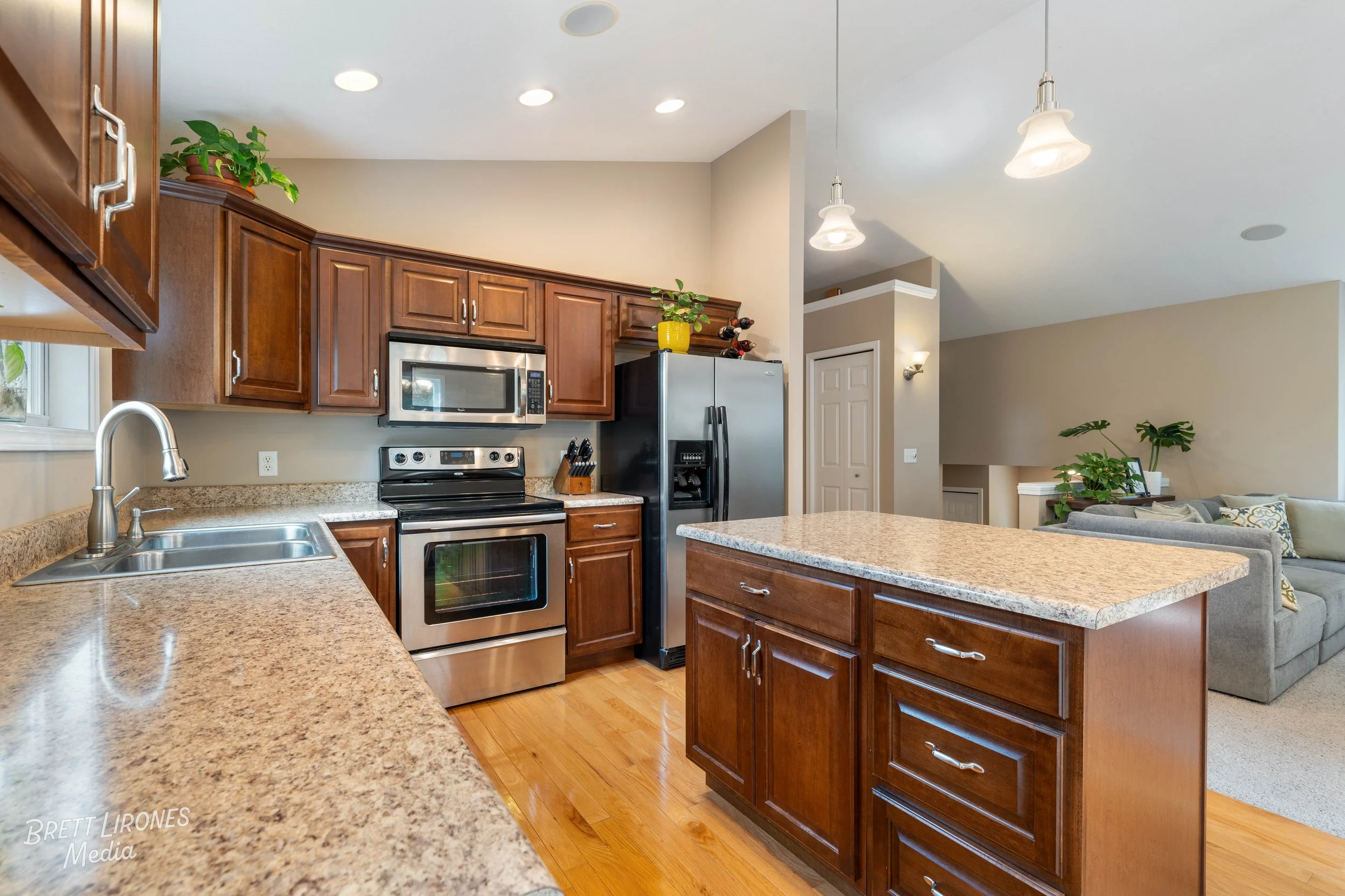 Kitchen with wooden cabinets, granite countertops, stainless steel appliances including a microwave, oven, and refrigerator, with a view into a living room with a gray sofa and plants.