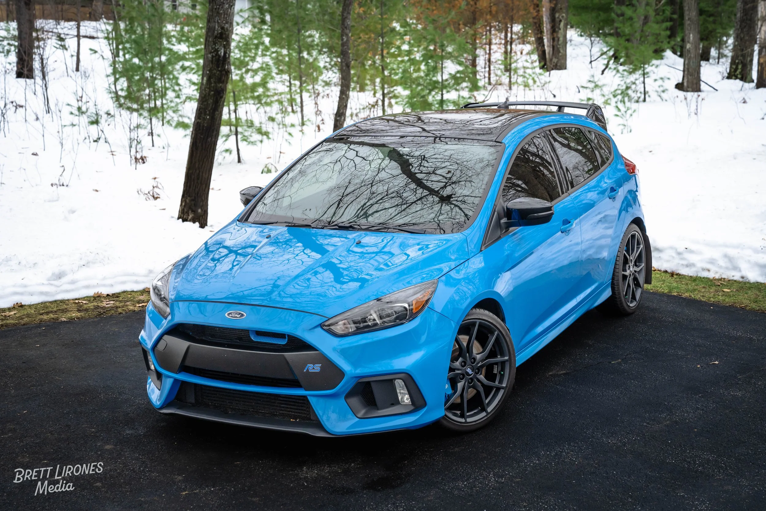A blue Ford Focus RS parked on a black pavement on a snowy background with a wooded area.