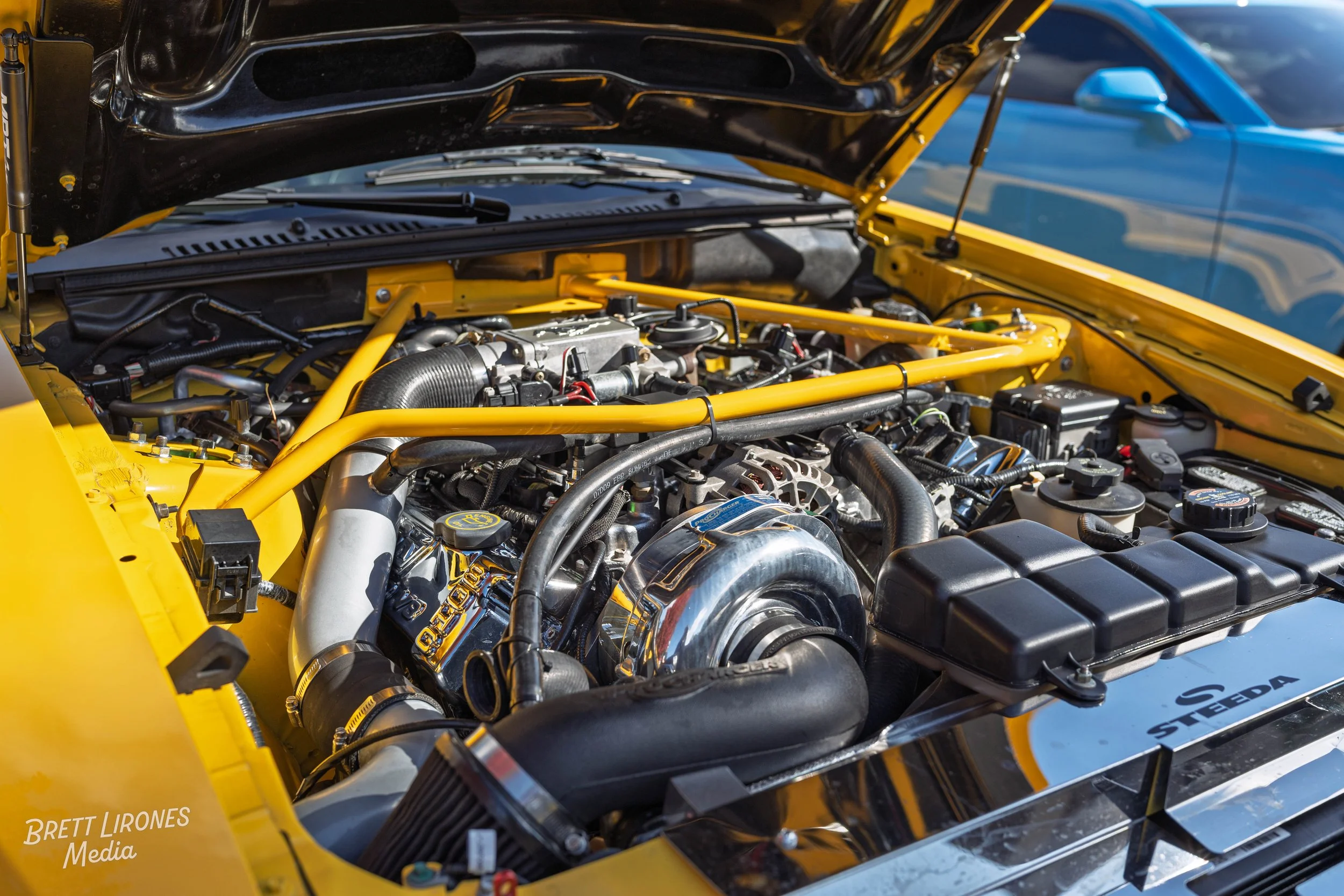 Close-up of a car engine bay with yellow accents and a polished alternator, showing various hoses, caps, and engine components inside the hood of a yellow car.