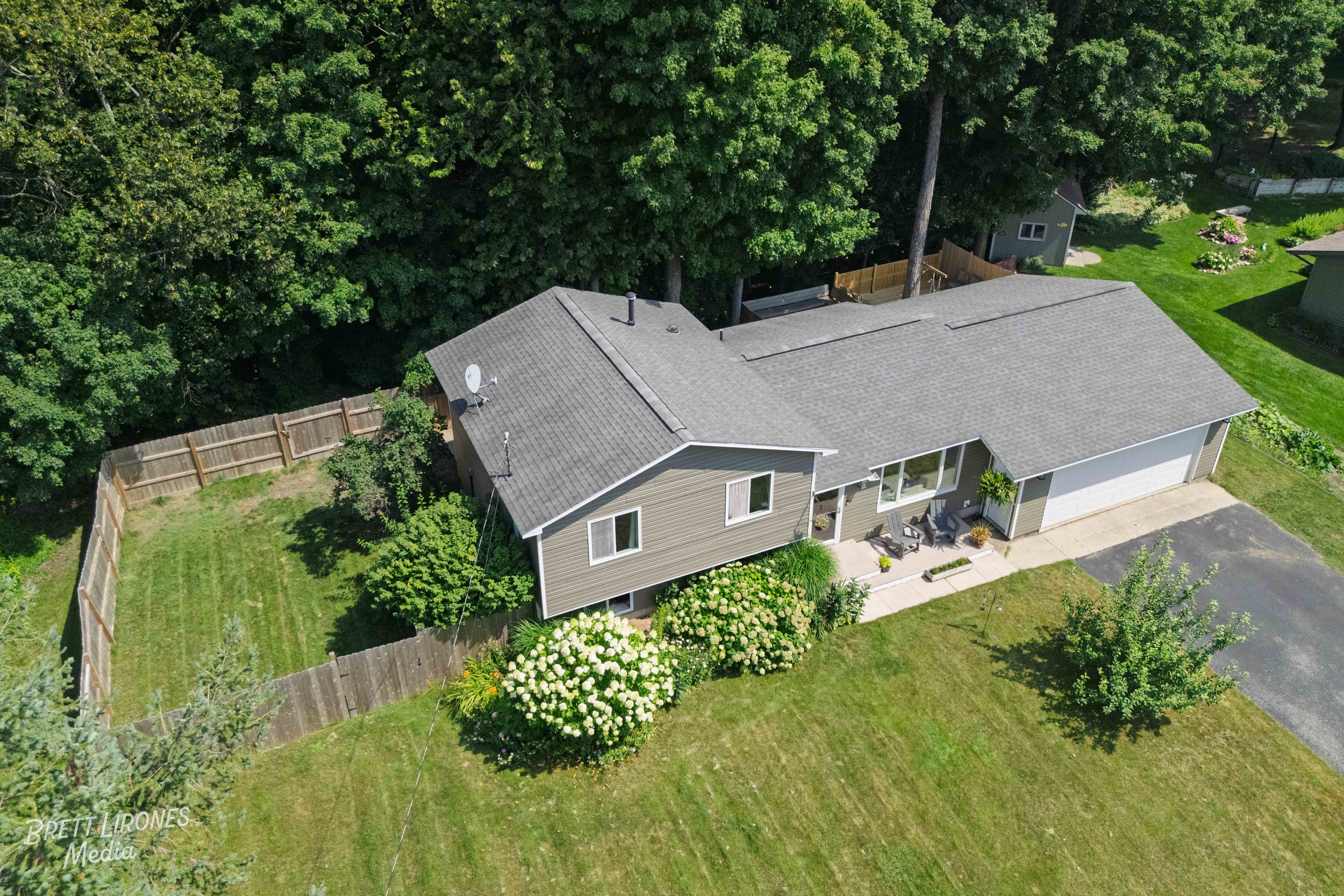 Aerial view of a single-story house with a gray shingle roof, surrounded by a green lawn and garden, with trees and neighboring houses nearby.