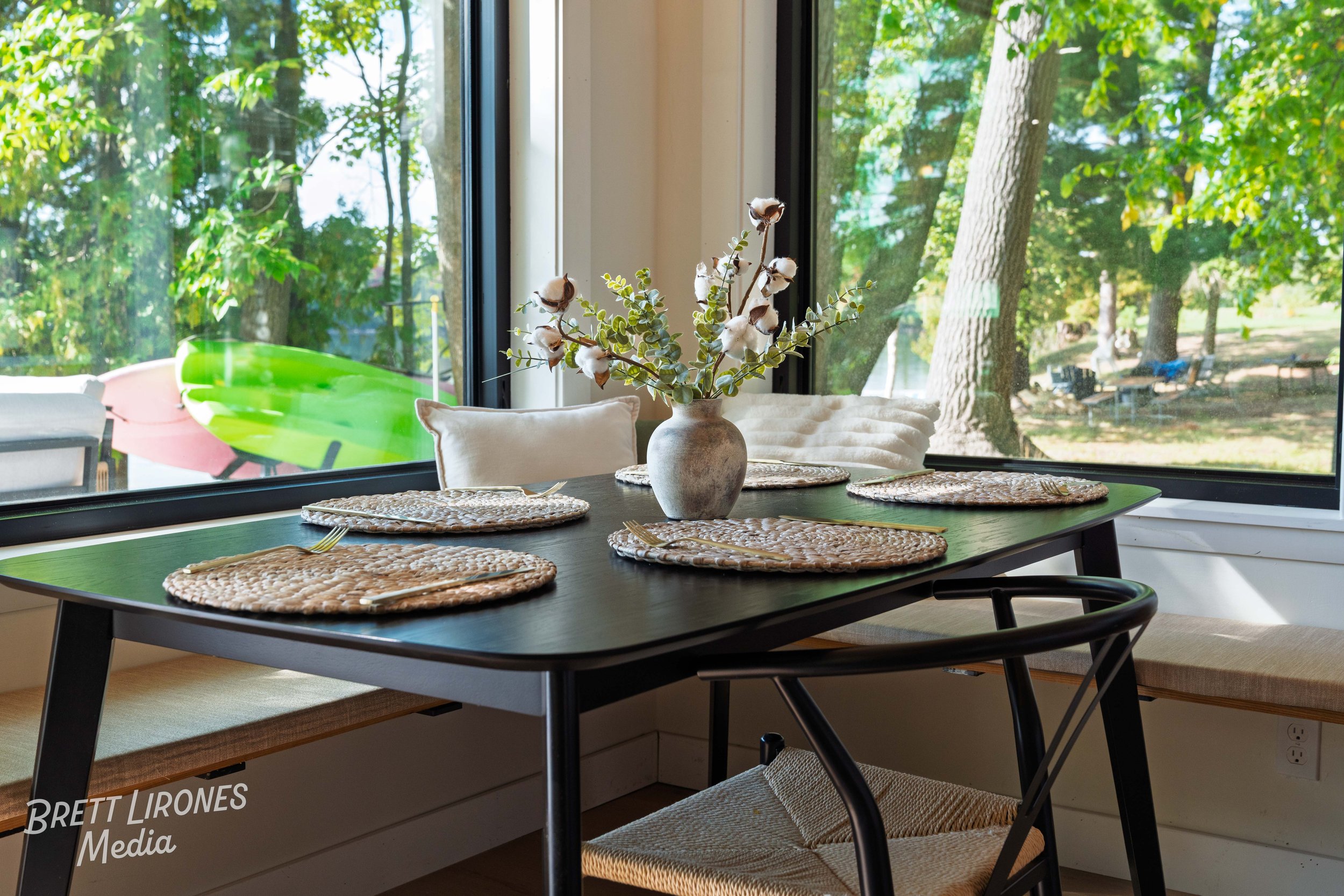 A dining table set near a large window with outdoor trees visible. The table has a vase with cotton and greenery, placemats, and utensils. Bench seating and cushions are around the table.